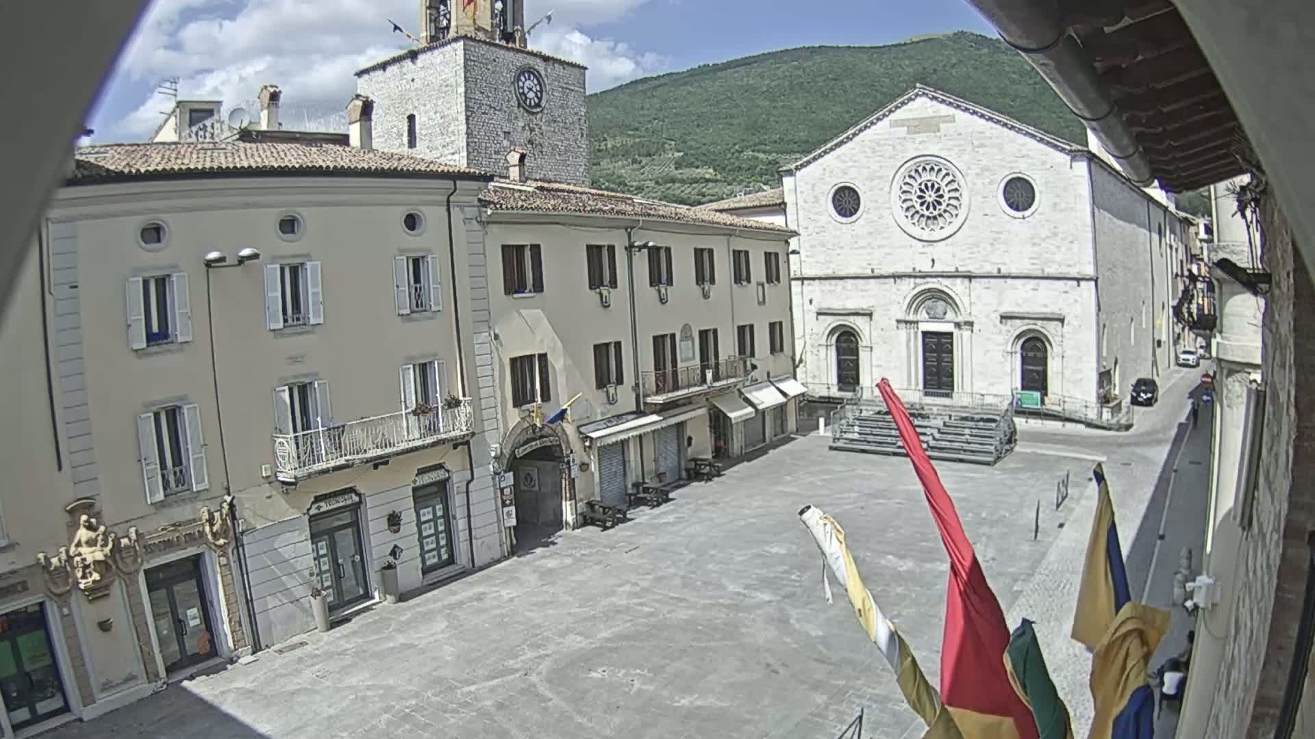 A town square under sunny skies is surrounded by buildings, including a church with a large rose window and a clock tower.