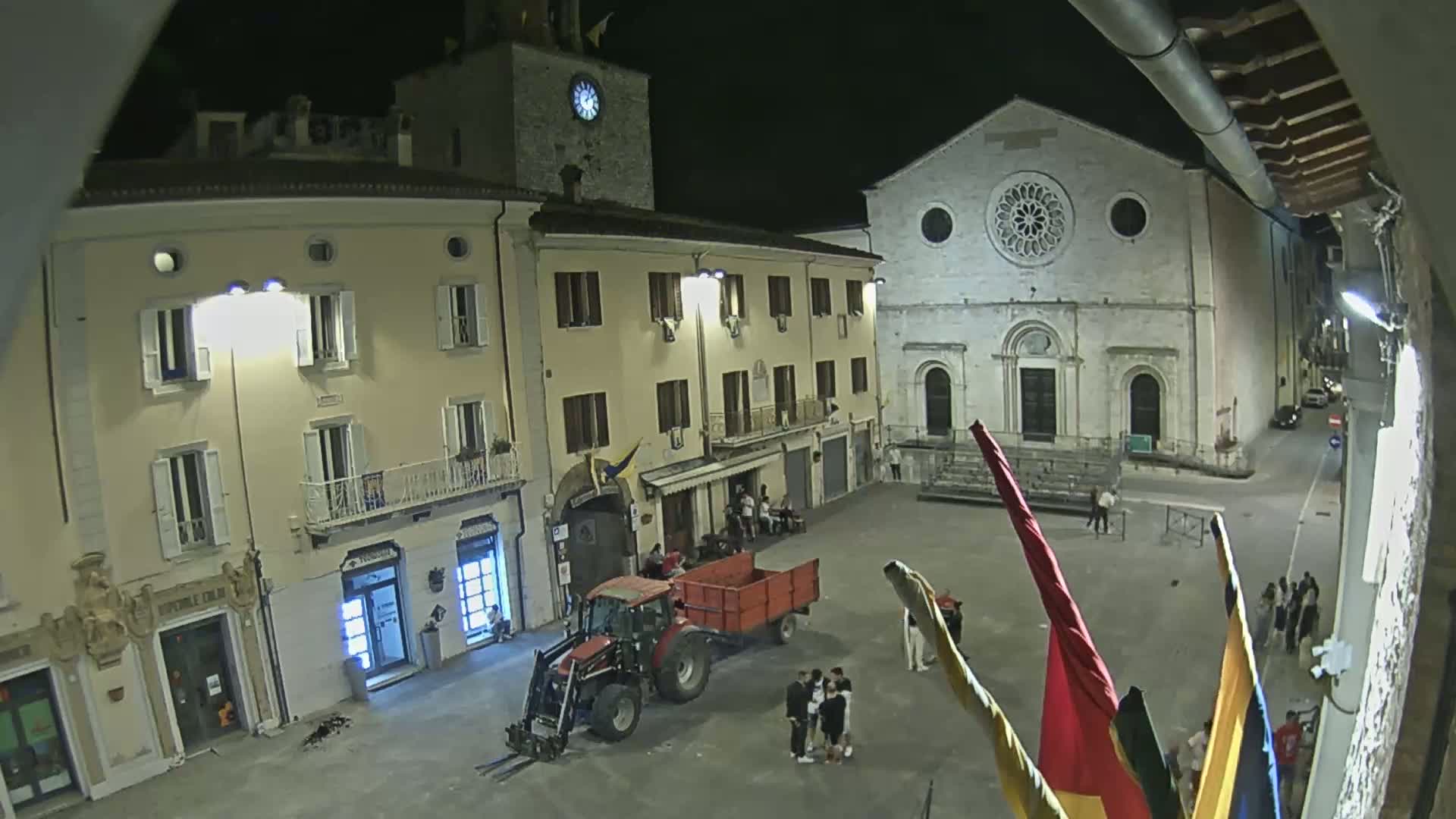 A nighttime, town square scene shows a tractor with a trailer parked near several people and flags, with buildings, including a church with a rose window, surrounding the square.