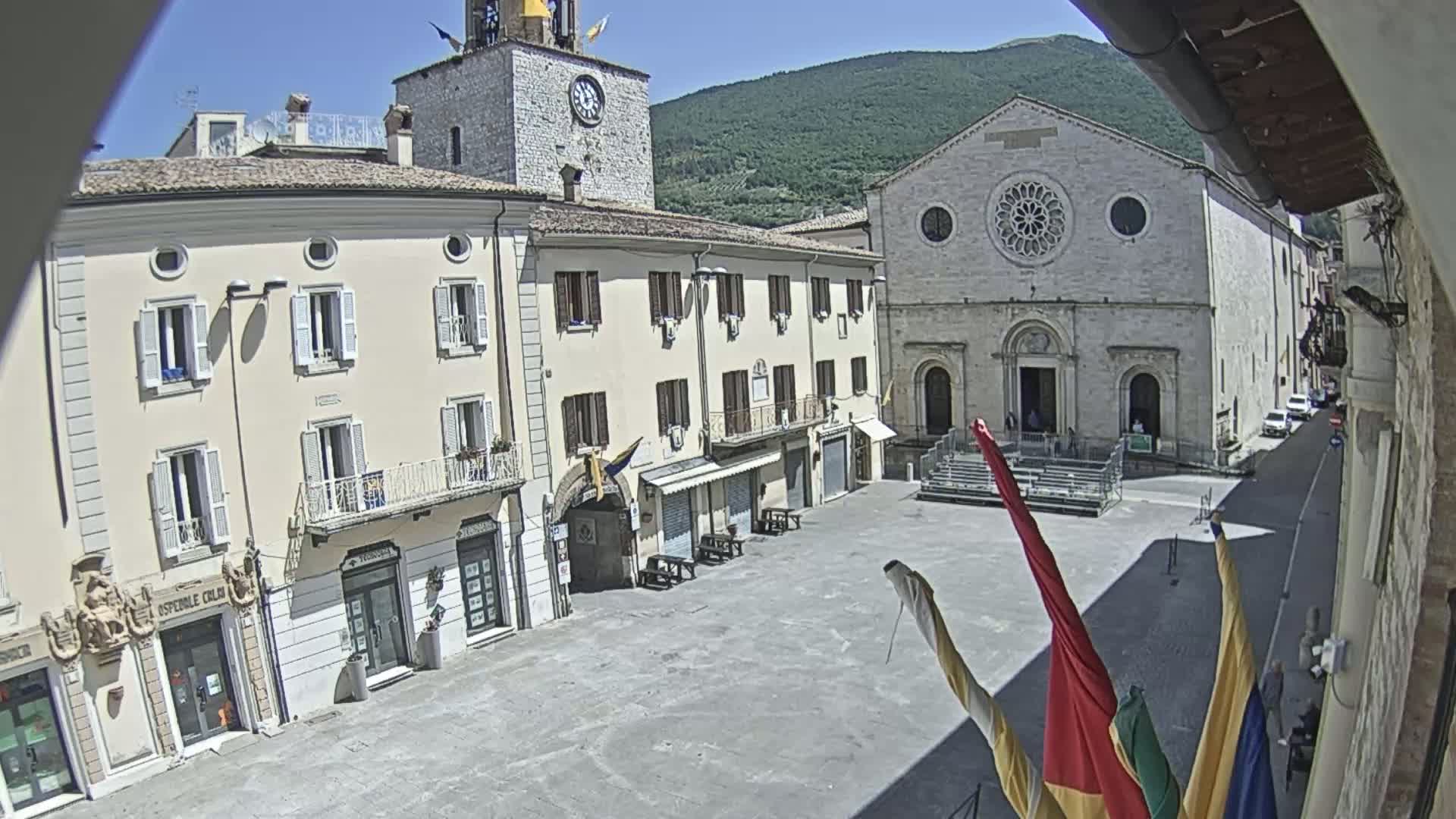 A sunny town square is surrounded by historic buildings, including a church with a rose window and a clock tower, under a clear sky and mountains in the background.