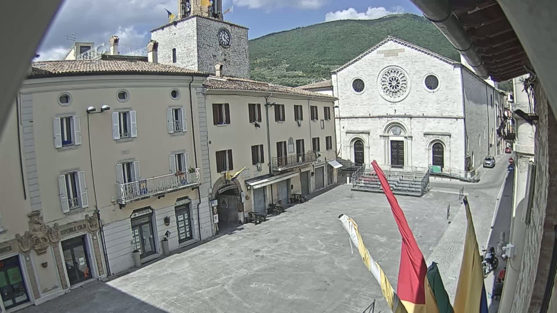 A town square with several buildings, including a church with a large rose window, under a sunny sky with mountains in the background.