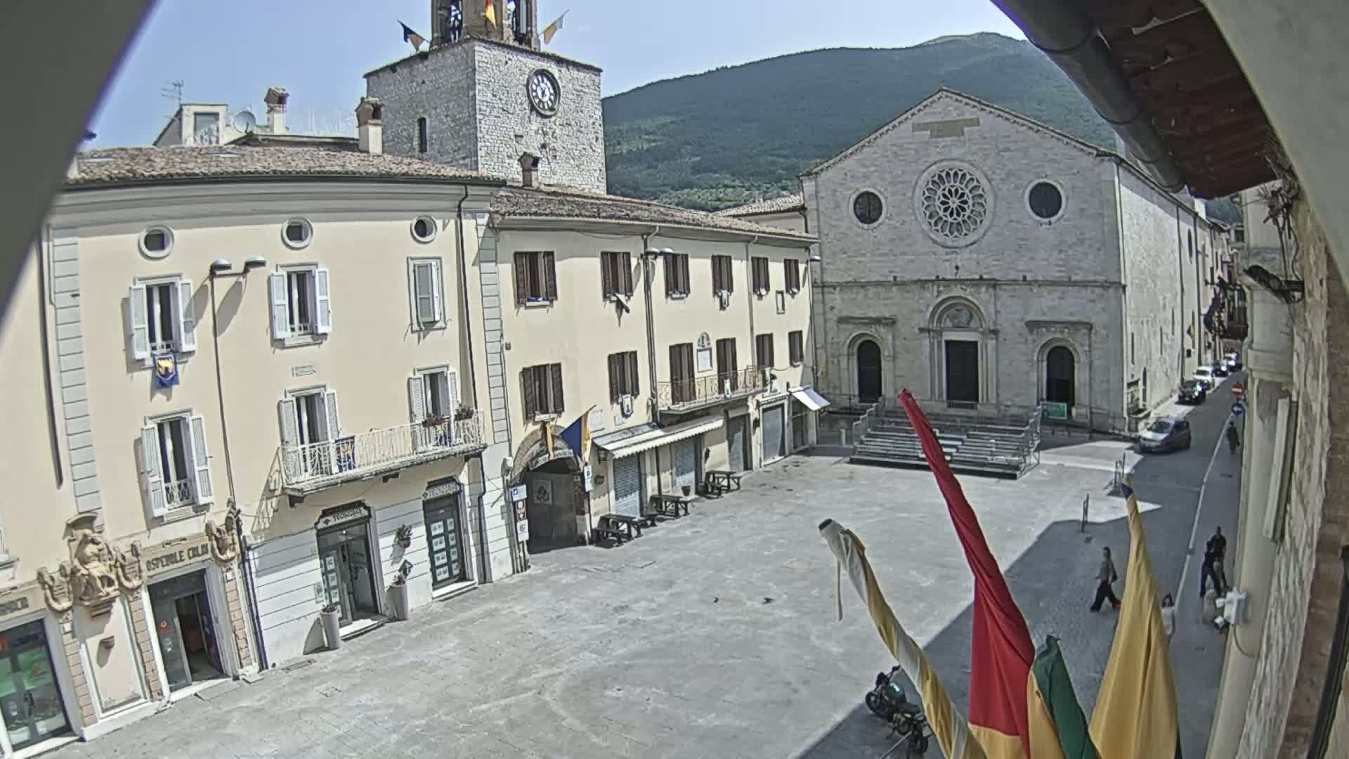 A town square with several buildings, including a church and clock tower, is viewed on a sunny day.