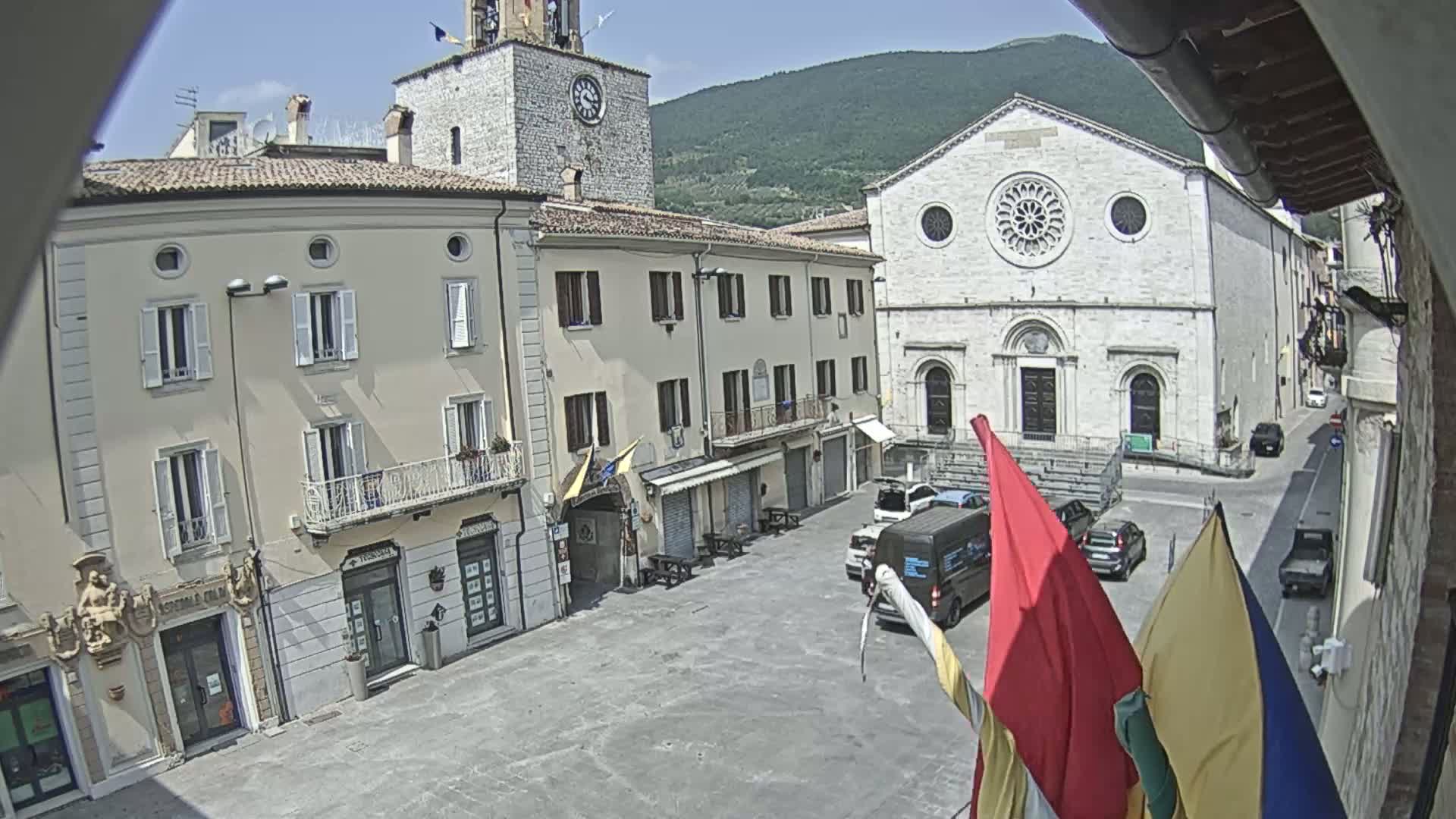 A sunny town square in a mountainous region features several buildings, including a church with a rose window, and a few cars are parked in the plaza.