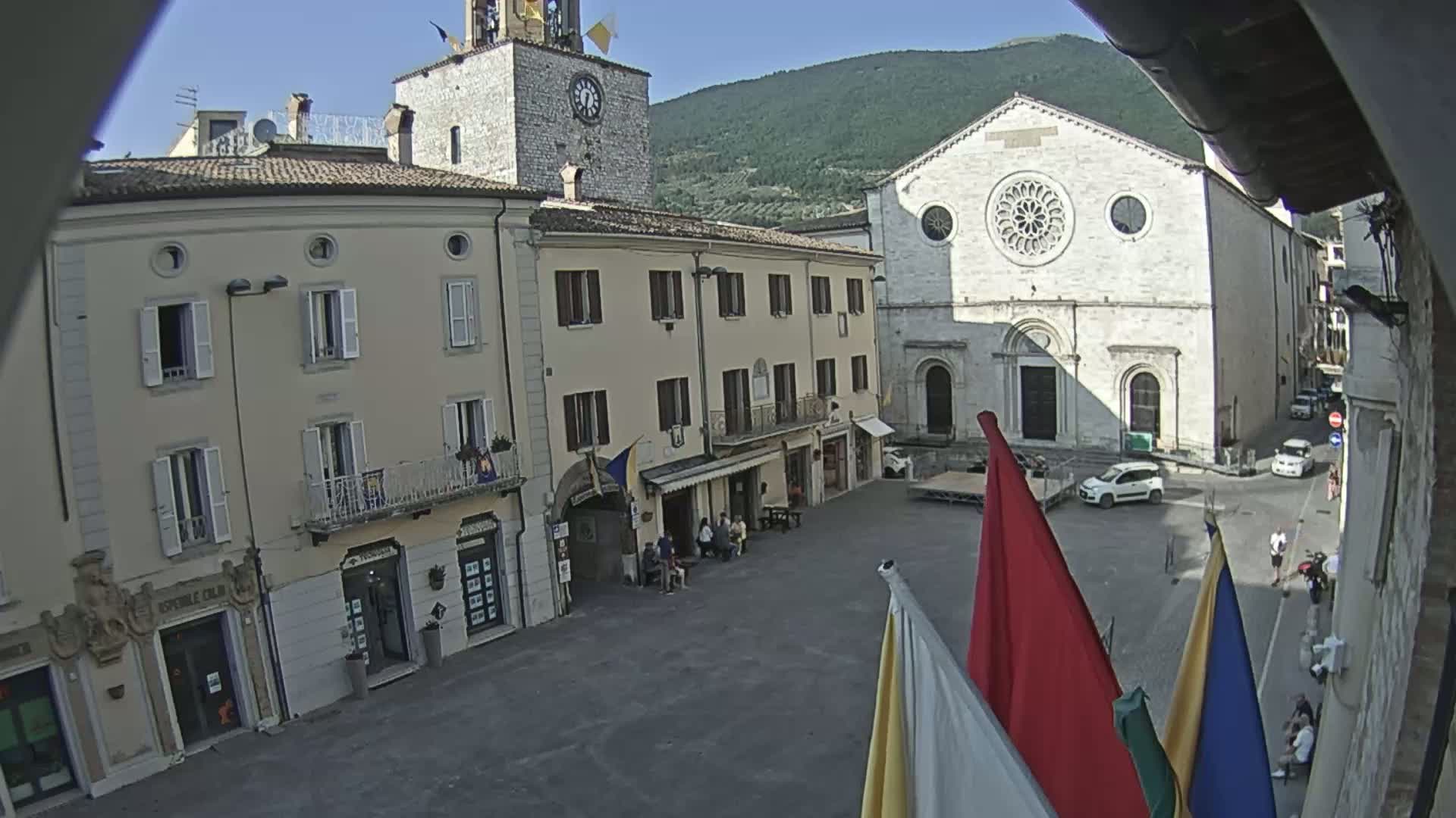 A town square with several buildings, including a clock tower and a large church, is seen on a clear, sunny day with a mountain in the background.