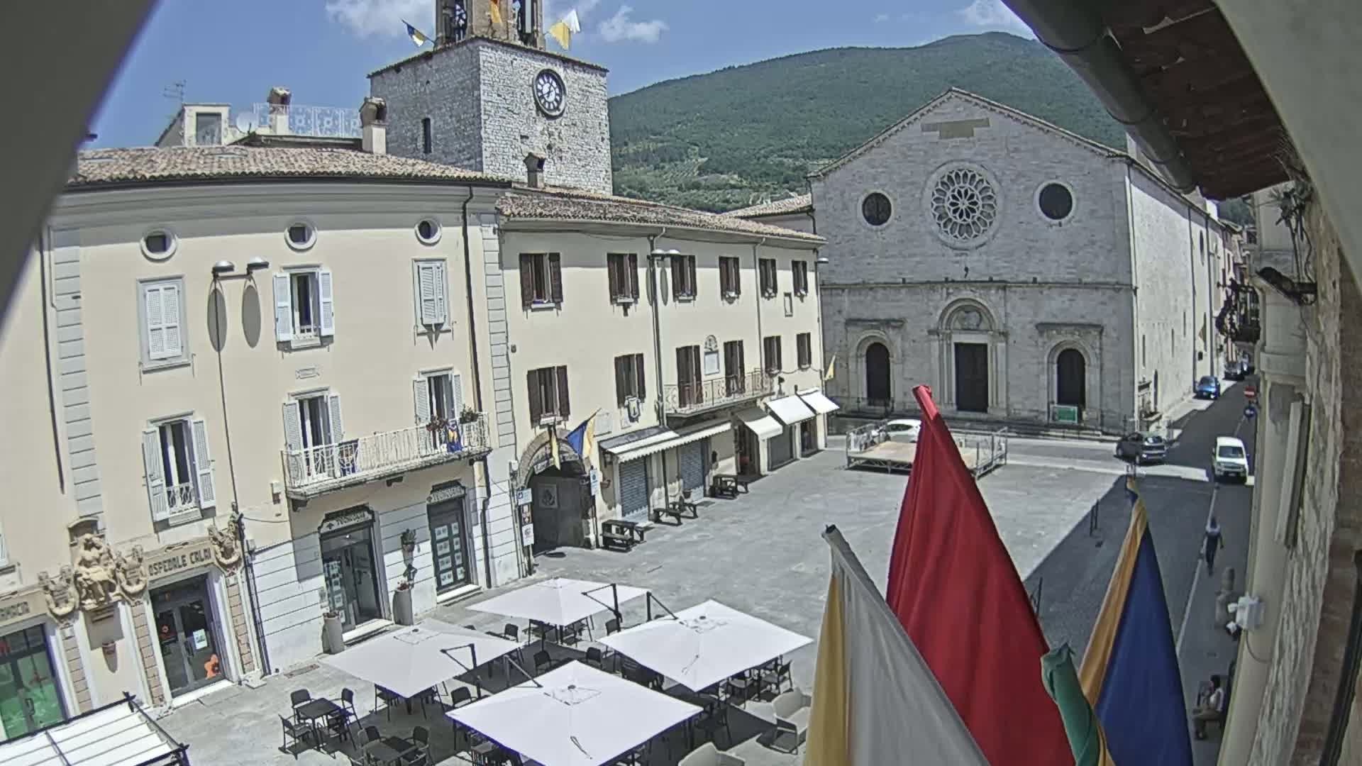 A sunny town square in Italy, surrounded by historical buildings including a clock tower and a church, features several outdoor cafe umbrellas and flags.