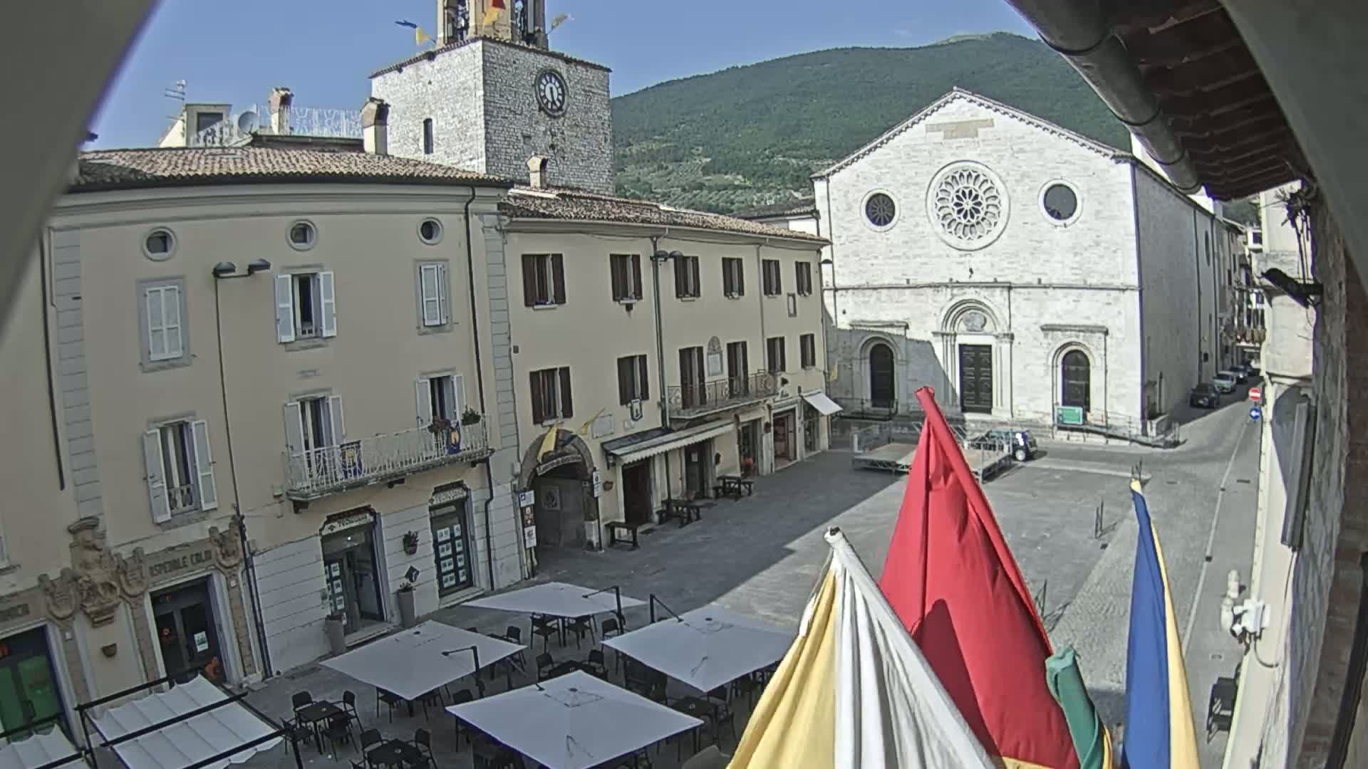 A sunny town square in Italy features several buildings, including a church with a rose window, a clock tower, and shops with outdoor seating.