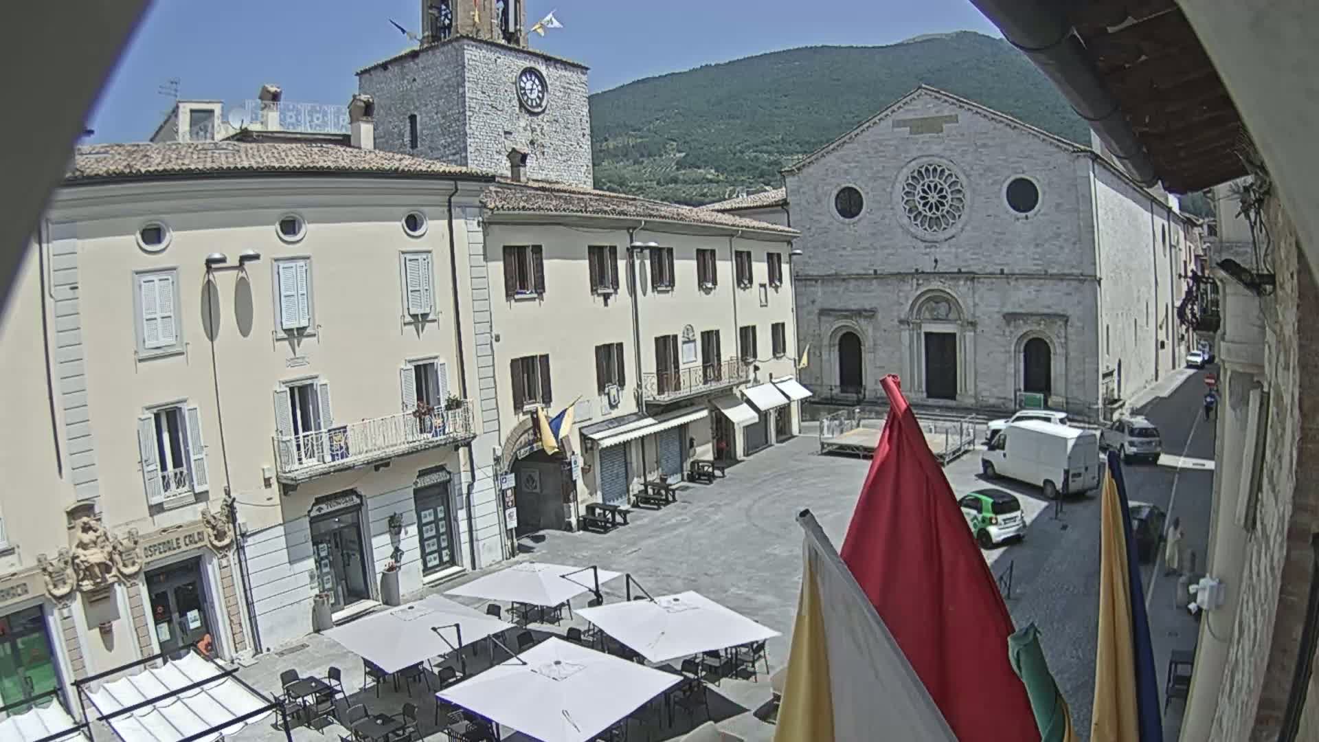 A sunny town square is shown, featuring several buildings, including a church with a rose window and a clock tower, with outdoor seating under umbrellas.