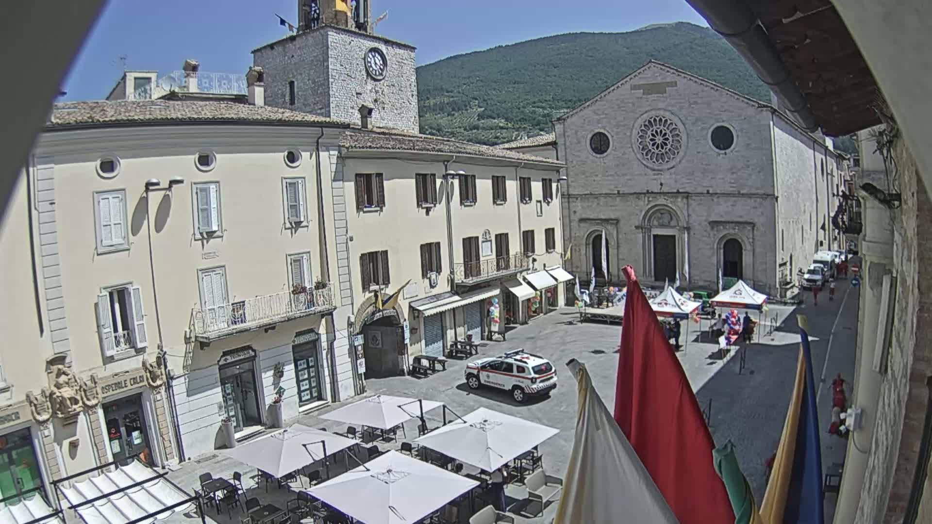 A sunny town square with several buildings, including a church and clock tower, features several market stalls and outdoor seating under umbrellas.