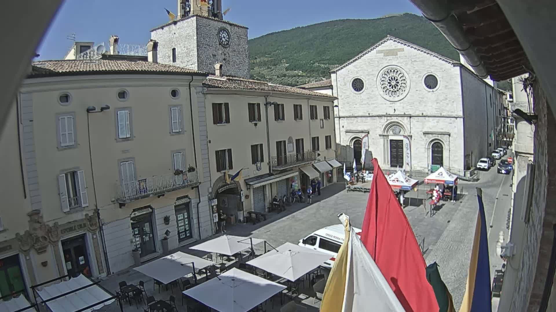 A sunny town square is shown, featuring several buildings including a church with a large rose window, a clock tower, and shops with outdoor seating, with some people and several flags visible.