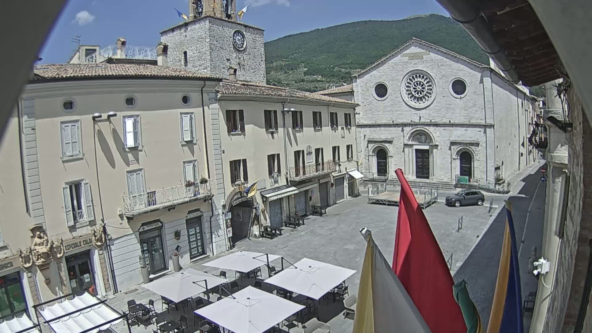A sunny town square, surrounded by historic buildings including a church with a rose window, features several outdoor cafe umbrellas and flags.