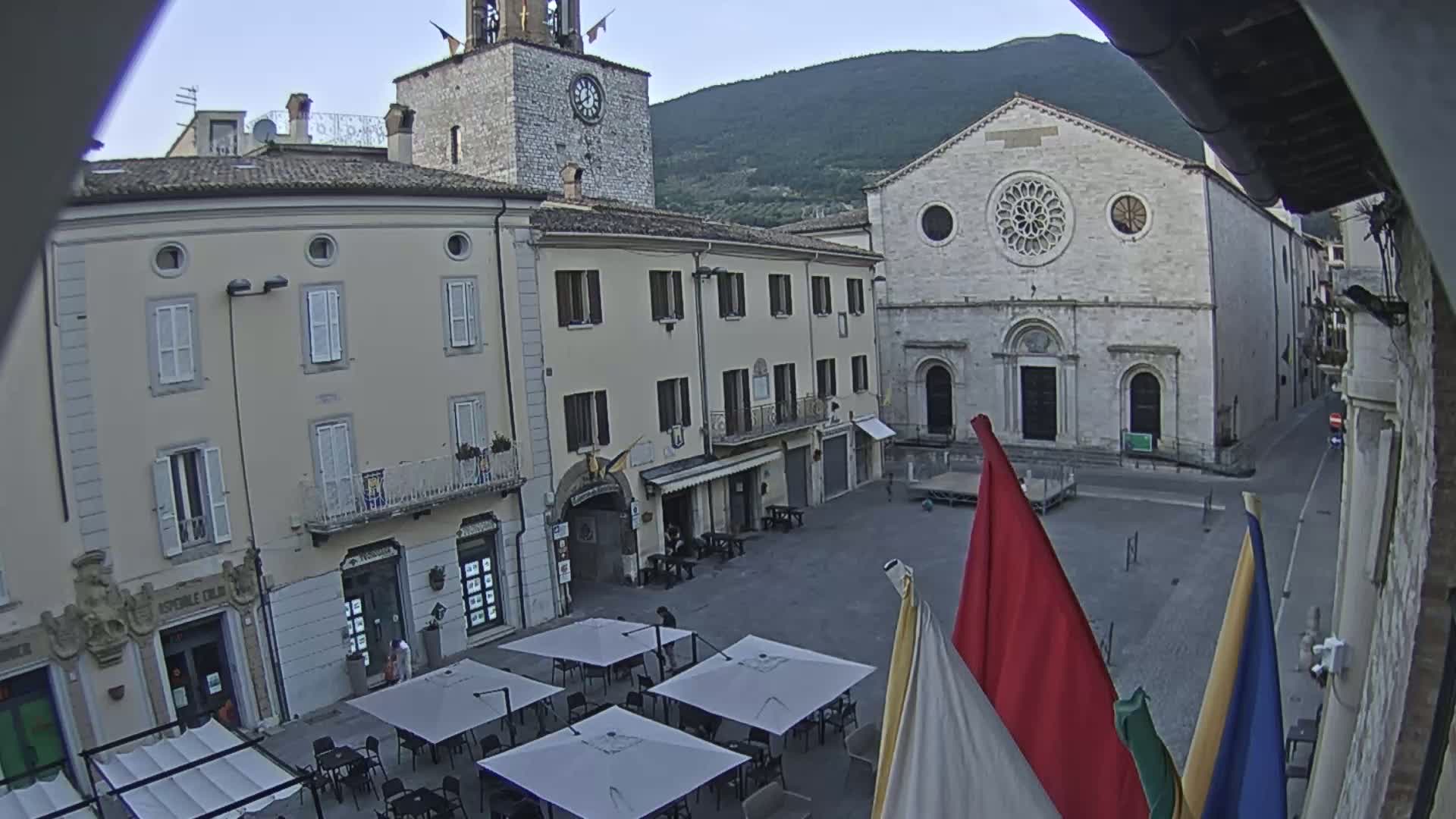 A town square with several buildings, including a clock tower and a church, is shown on an overcast day with several outdoor tables under umbrellas.