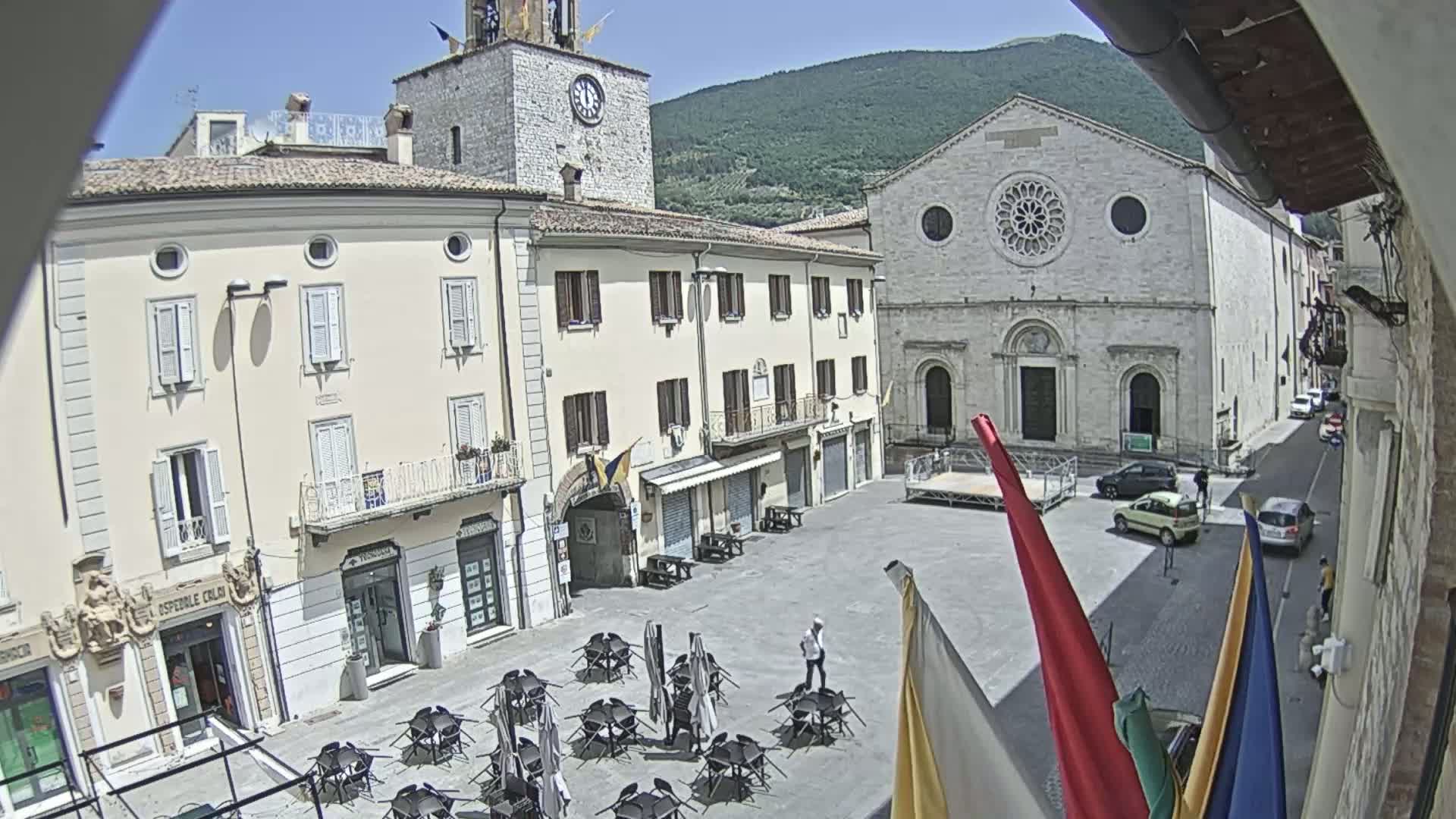A sunny town square, partially surrounded by buildings, including a clock tower and a large church, with several tables and chairs set up outdoors.