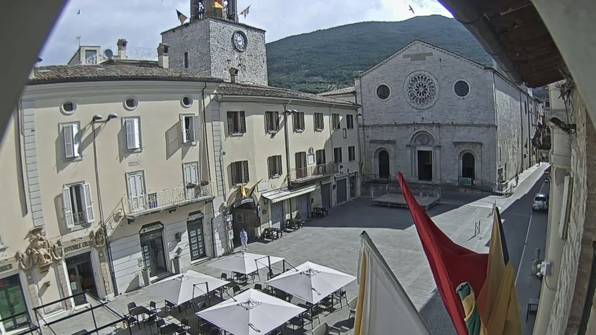 A sunny view of a town square with several buildings, including a clock tower and a church, with outdoor seating under umbrellas.