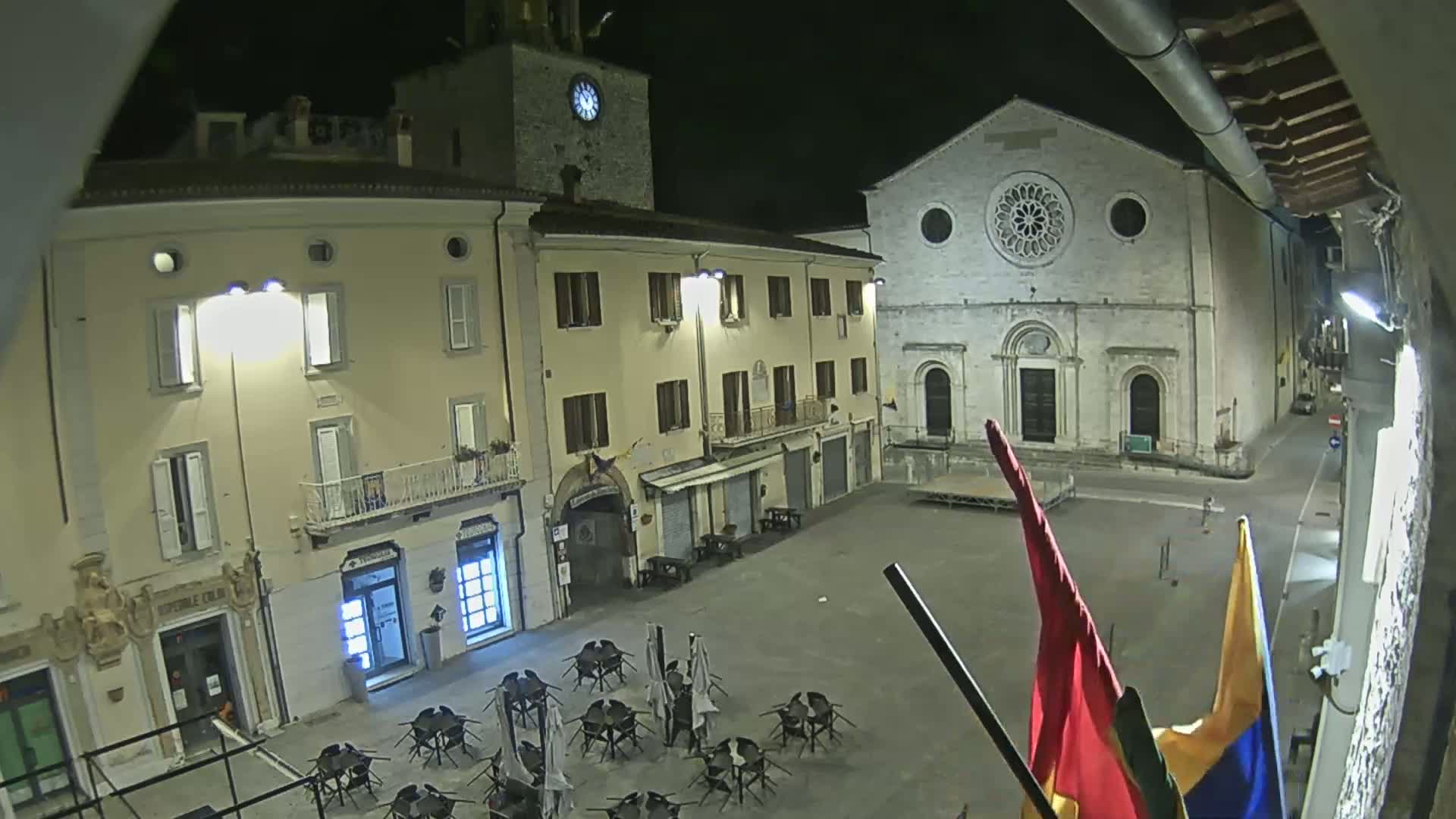 A nighttime view of a town square with several buildings, including a church with a large rose window, and outdoor seating in the square under nighttime lighting.
