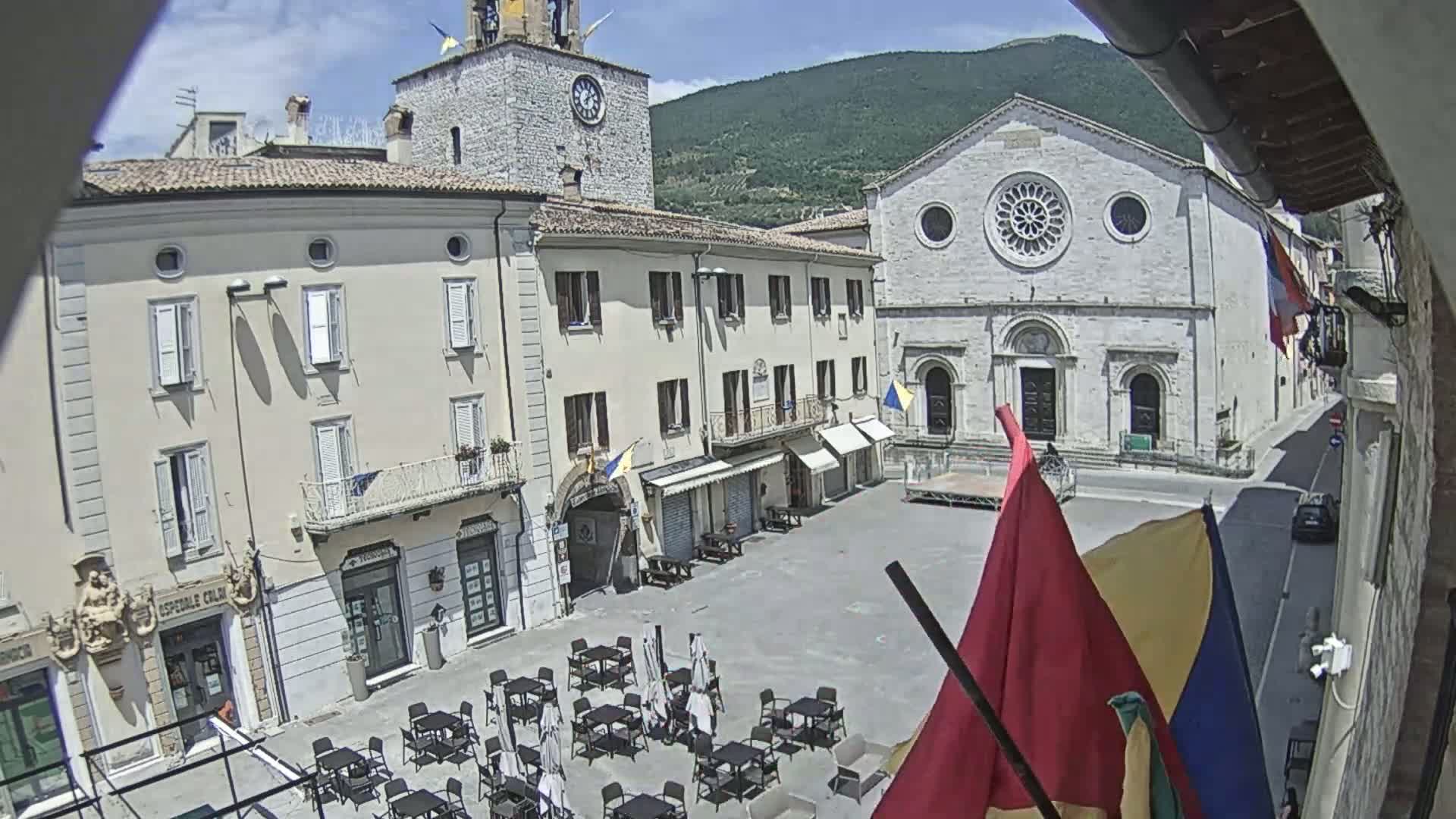 A sunny town square with several buildings, outdoor seating, and flags visible, backed by a mountain range.