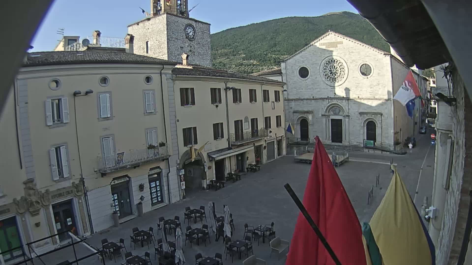 A town square with several buildings, including a church and clock tower, is viewed on a clear day with a mountain in the background.