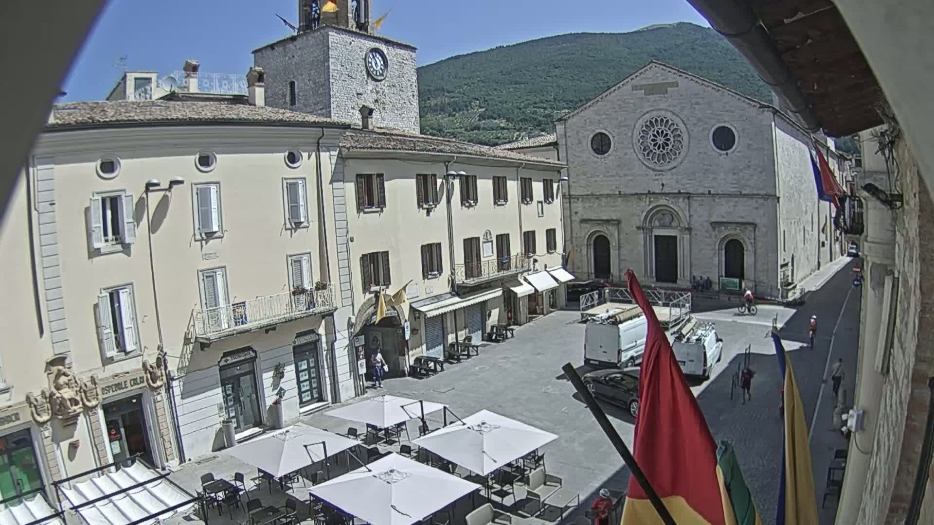 A sunny town square is shown, featuring several buildings, including a church with a rose window, a clock tower, and outdoor café seating.