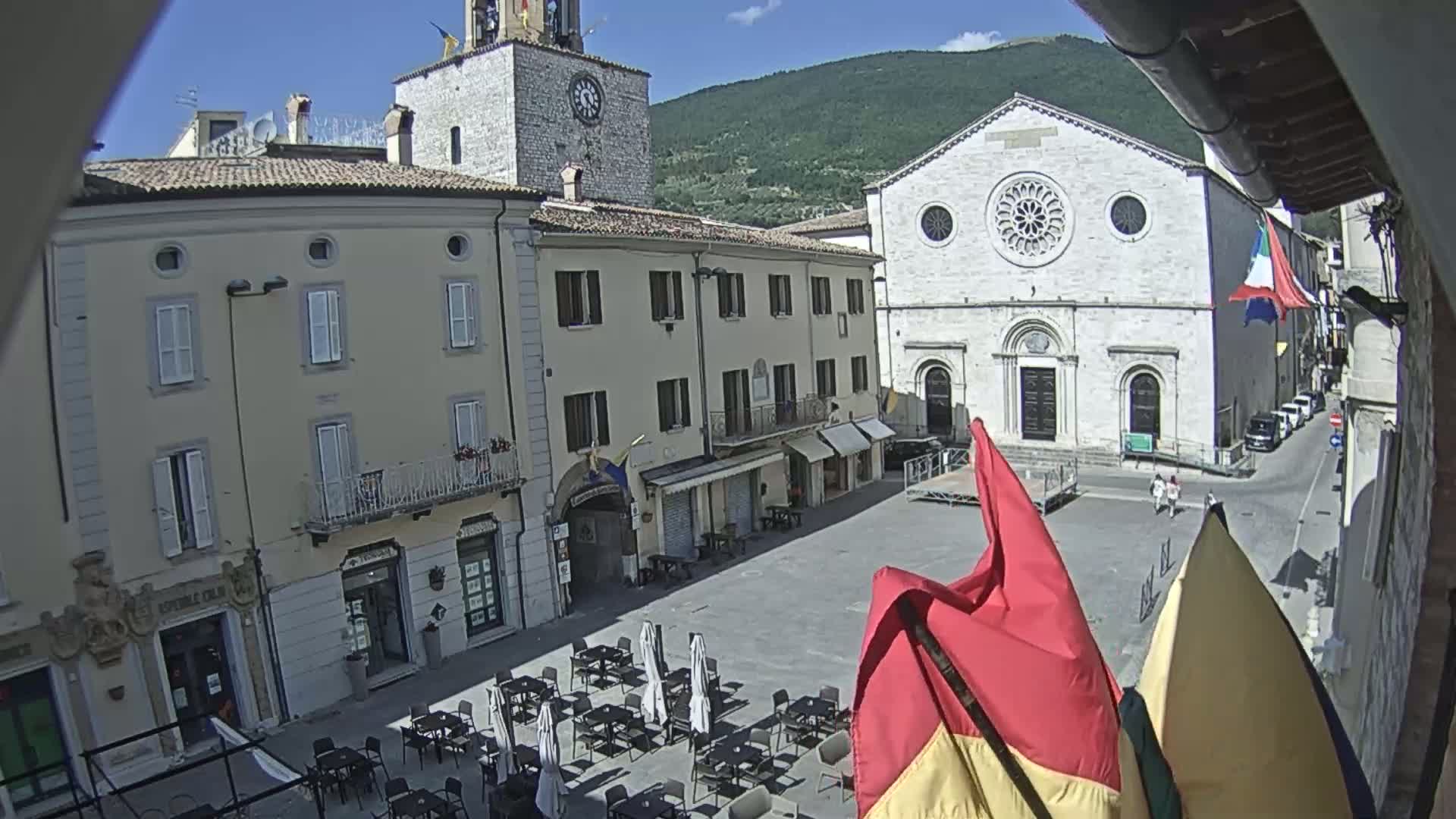 A town square with several buildings, including a church with a rose window, is seen on a sunny day with a mountain in the background.