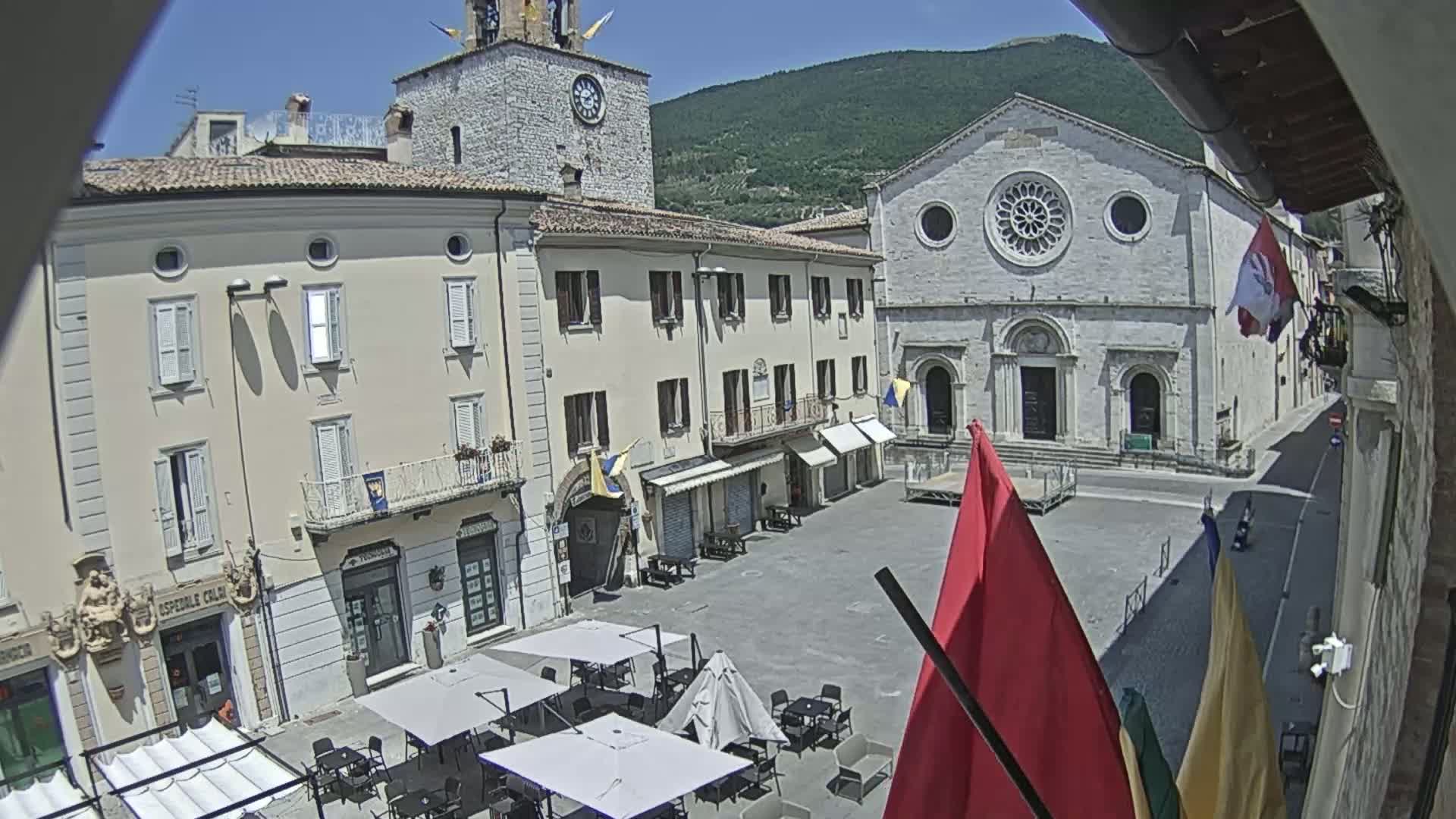 A town square, under sunny skies, is surrounded by light-colored buildings, including a church with a large rose window, and contains several outdoor café umbrellas and flags.