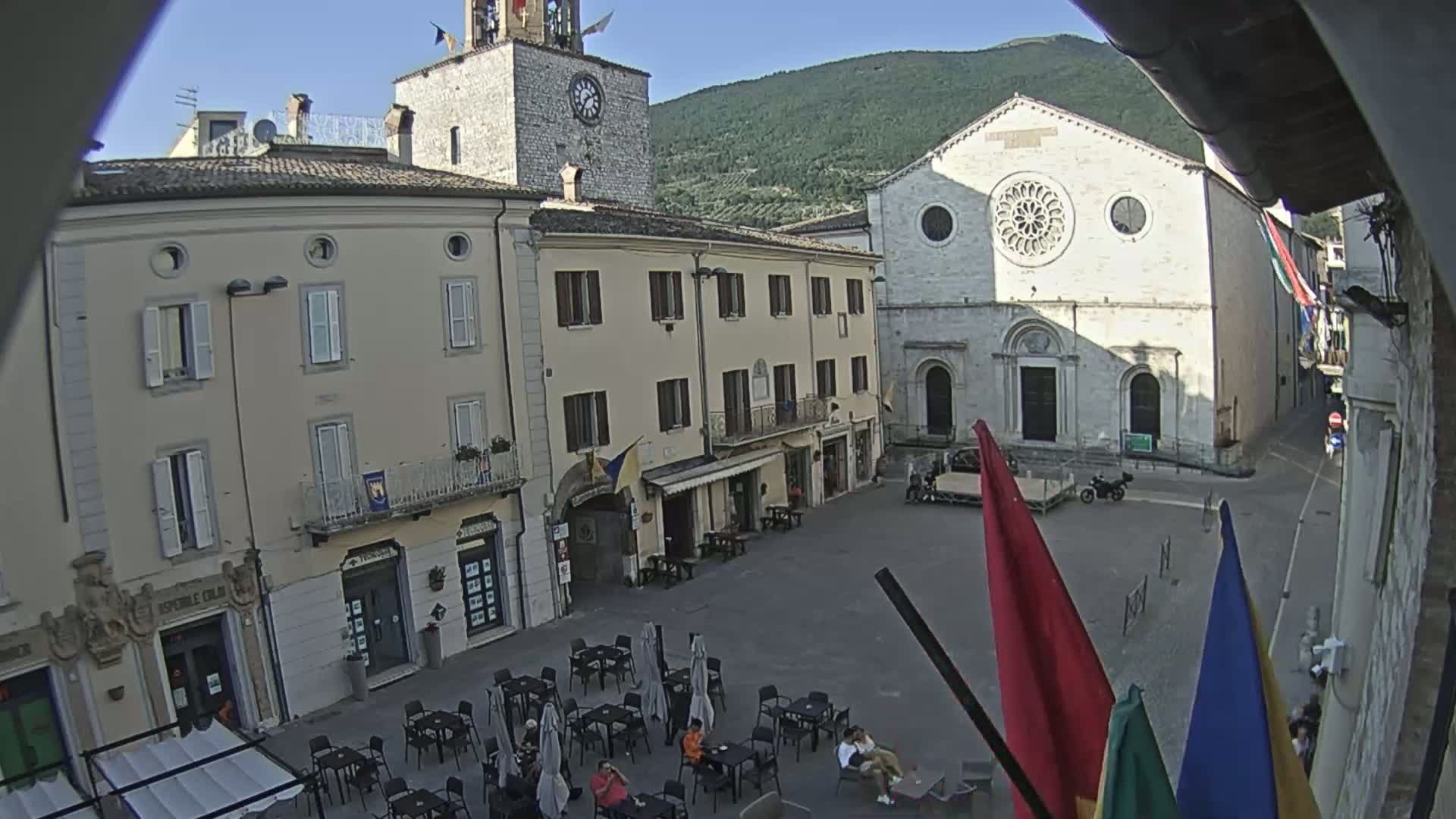 A town square with several buildings, including a church with a rose window, is seen on a sunny day, with a few people seated at outdoor tables.
