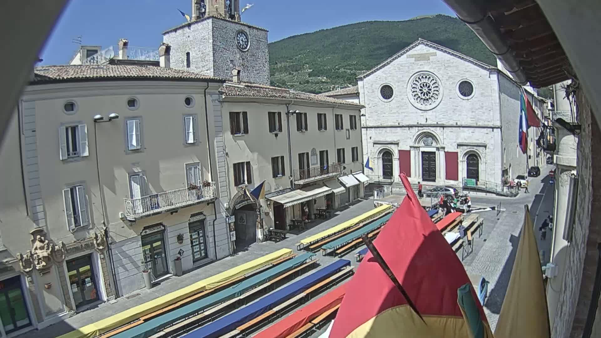 A sunny town square features numerous long tables set up, flanked by buildings, one of which is a church with a large rose window, all under a clear sky with mountains in the background.