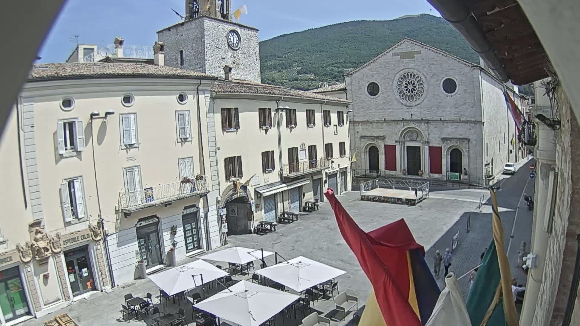 A sunny town square with several buildings, including a church with a rose window, is partially visible from a high vantage point, with several outdoor café umbrellas set up.