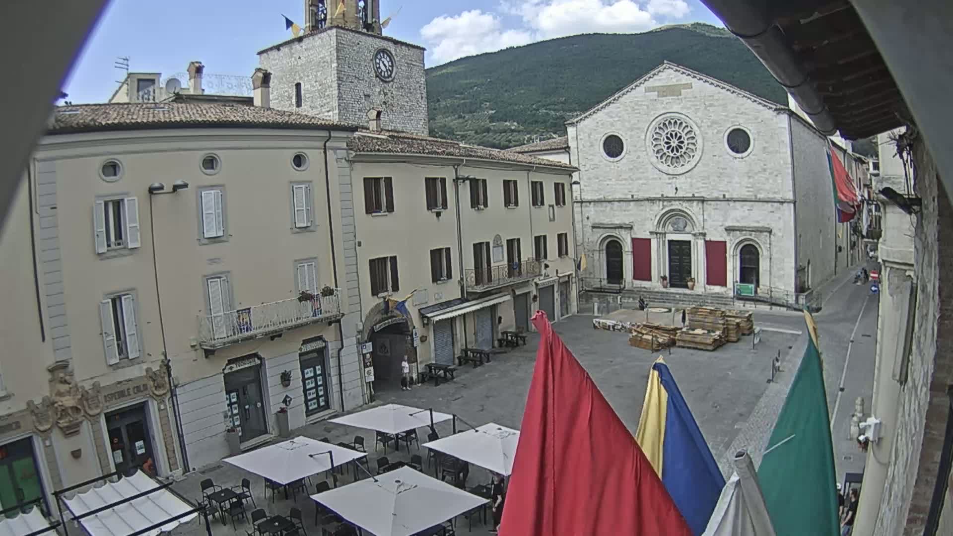 A town square with several buildings, including a church with a rose window, is partially obscured by flags on a partly cloudy day.