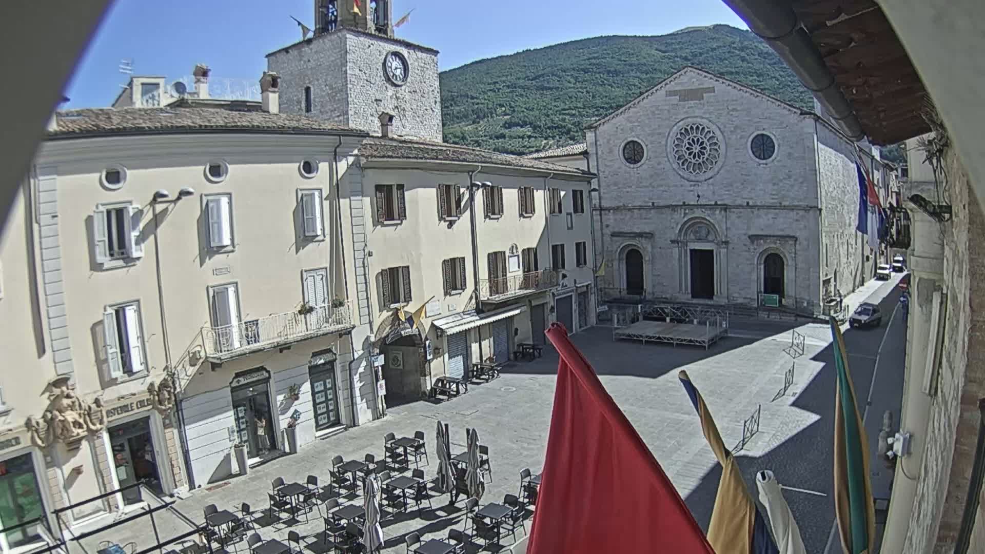 A sunny outdoor view of a town square with several buildings, including a church and a clock tower, with a few cars parked on a side street, and outdoor cafe seating.