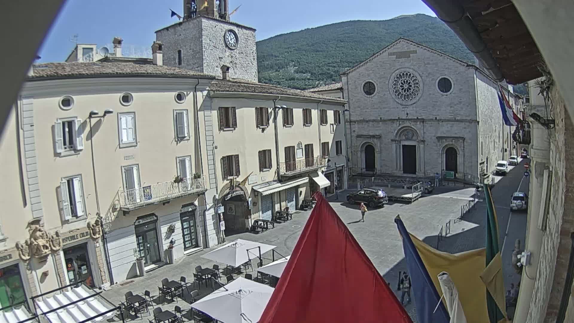 A sunny town square is viewed from a balcony, showing several buildings, a church with a rose window, outdoor seating, and a few cars parked nearby.