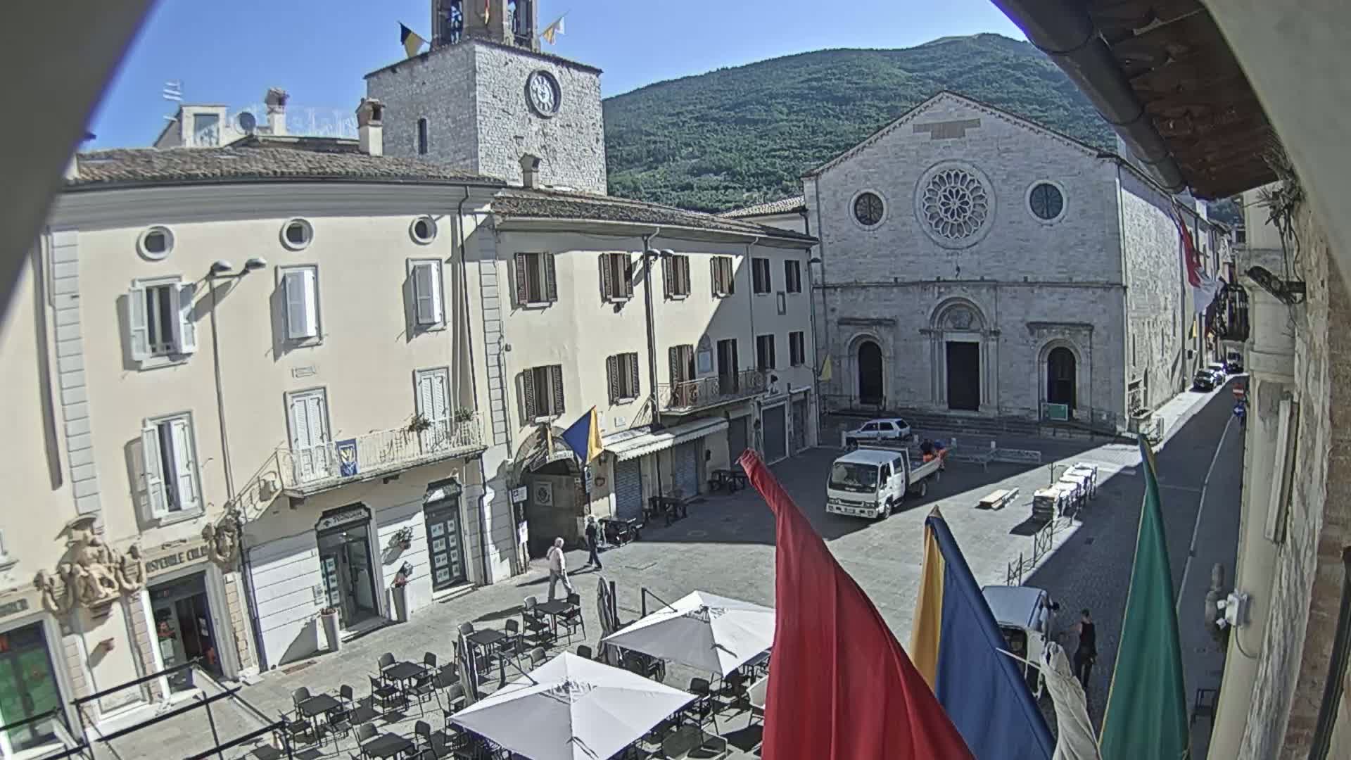 A sunny town square, surrounded by historic buildings, including a church with a rose window and a clock tower, features outdoor seating and a few people.