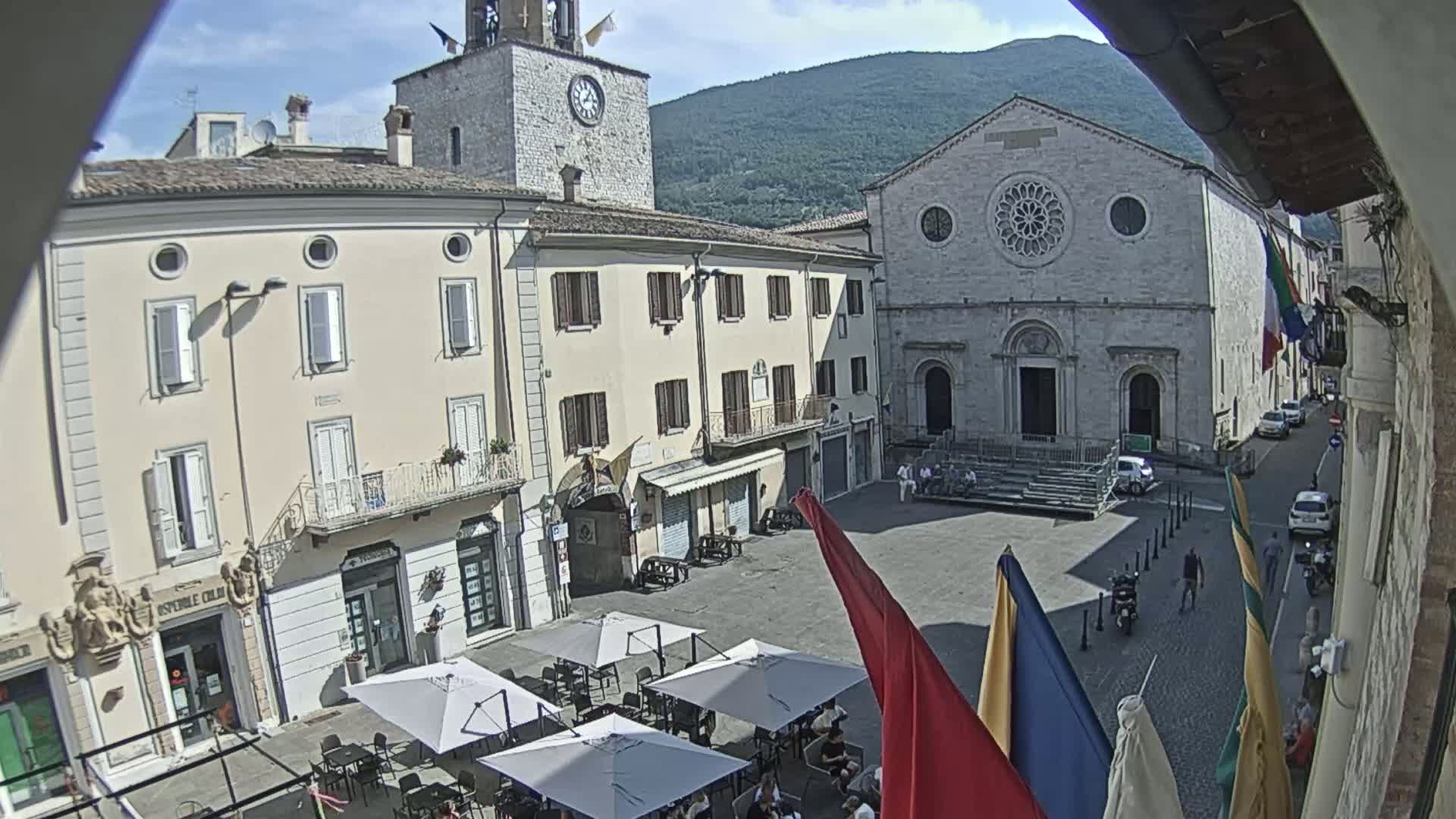 A sunny town square with outdoor seating, a clock tower, and a large stone church is backed by a mountain under a mostly clear sky.