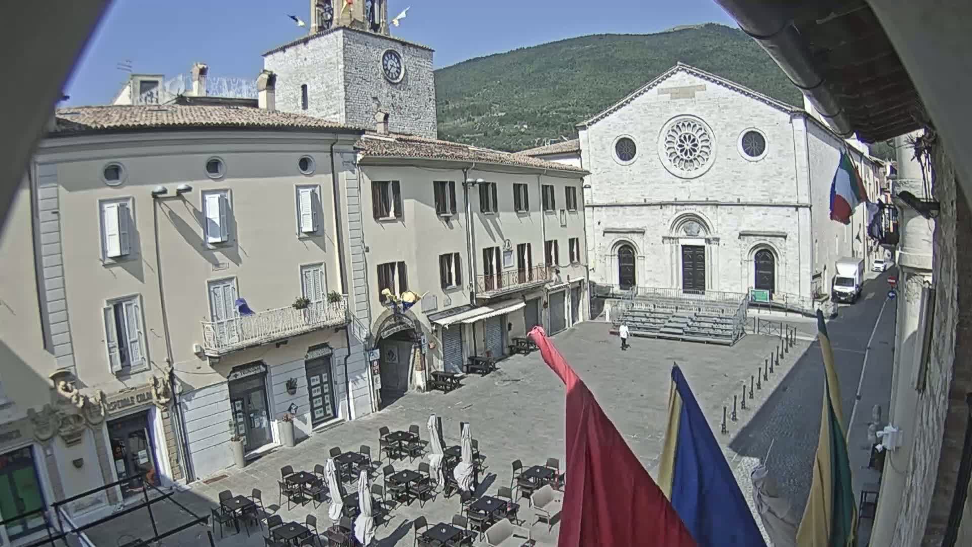 A sunny town square with outdoor seating, a clock tower, and a large stone church is backed by a mountain under a mostly clear sky.