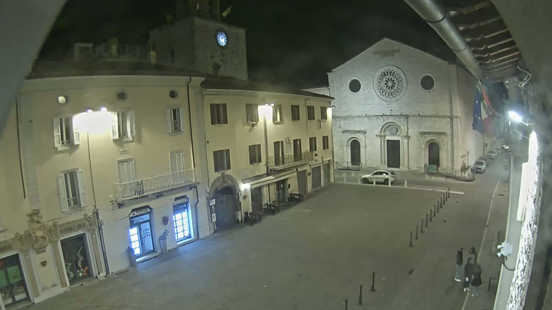 An illuminated town square at night, under clear skies, features a large stone church with a rose window, a prominent clock tower, multi-story buildings with ground-floor shops, a parked white car, and a small group of people standing on the paved square.