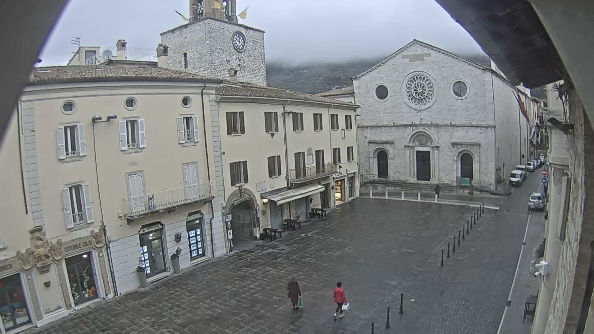 An overcast day reveals a wet, paved town square with two people walking, flanked by traditional multi-story buildings and a large stone church, with a clock tower and misty mountains in the background.