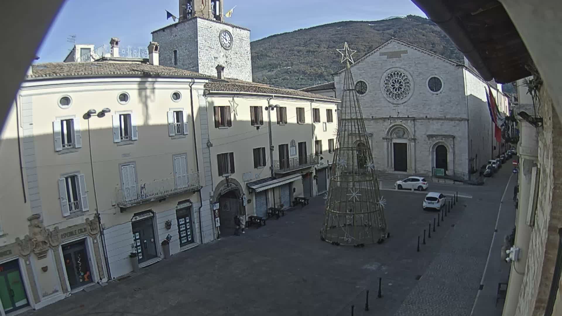 A sunny day illuminates a European town square featuring an unlit, decorative Christmas tree frame, a grand church with a rose window, a prominent clock tower, and surrounding multi-story buildings, all set against a backdrop of distant mountains.