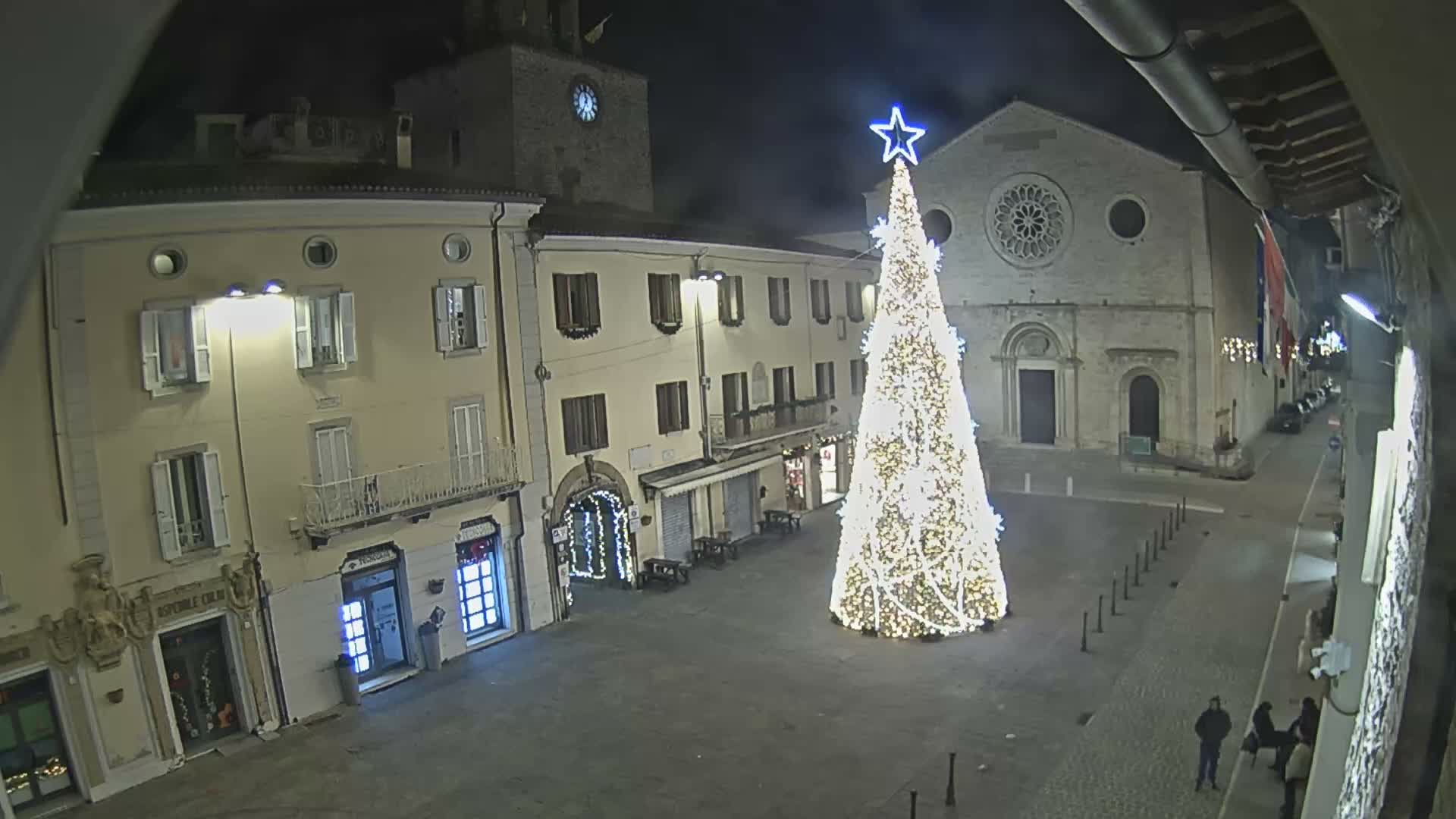 A large, brightly lit Christmas tree with a blue star adorns a historic European town square at night, surrounded by illuminated multi-story buildings and a stone church, with a few people scattered around under clear conditions.
