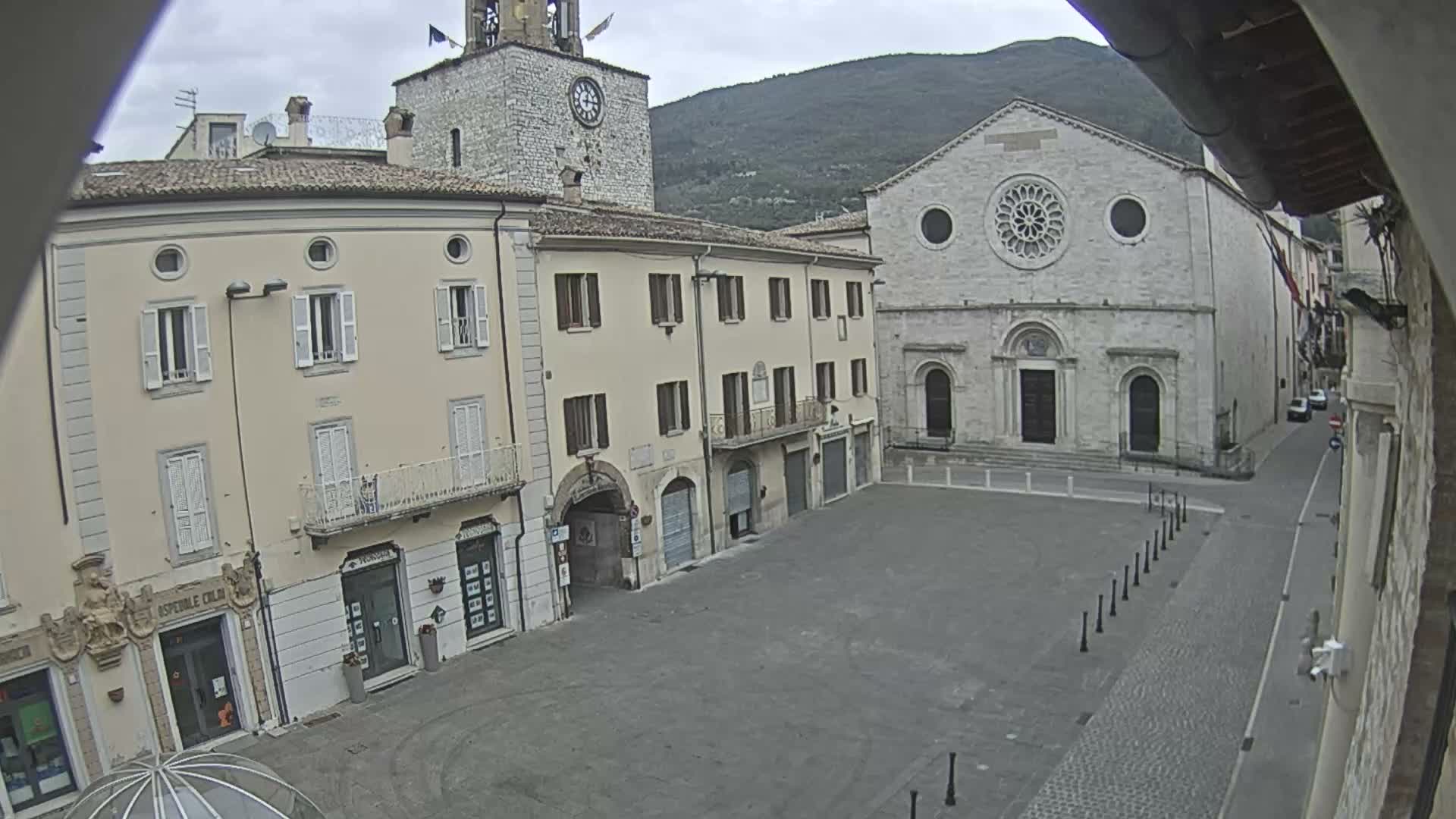 An overcast day reveals a European town square adorned with a large illuminated Christmas tree, flanked by historic stone buildings including a church and a clock tower, with distant mountains under a gray sky.