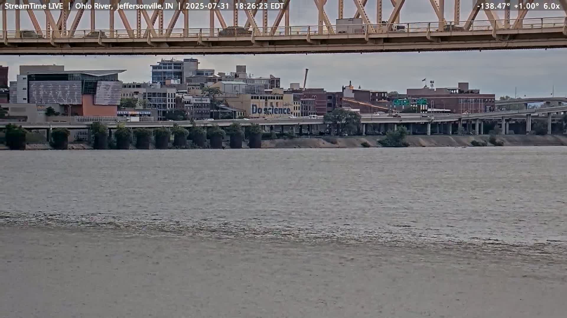 A river flows under a bridge, with a city skyline and overcast skies visible in the background.