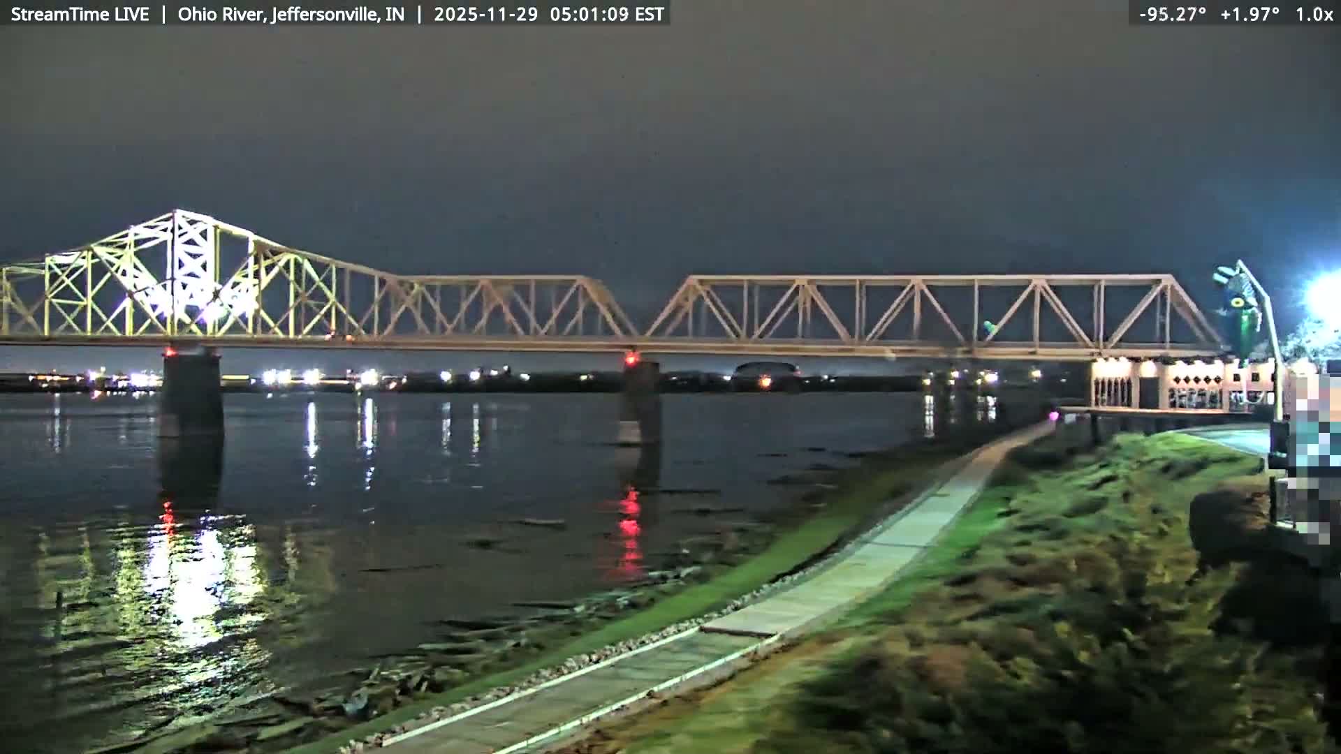 An illuminated truss bridge stretches across a dark river under an overcast night sky, with lights reflecting on the water and a paved pathway bordering the right bank.