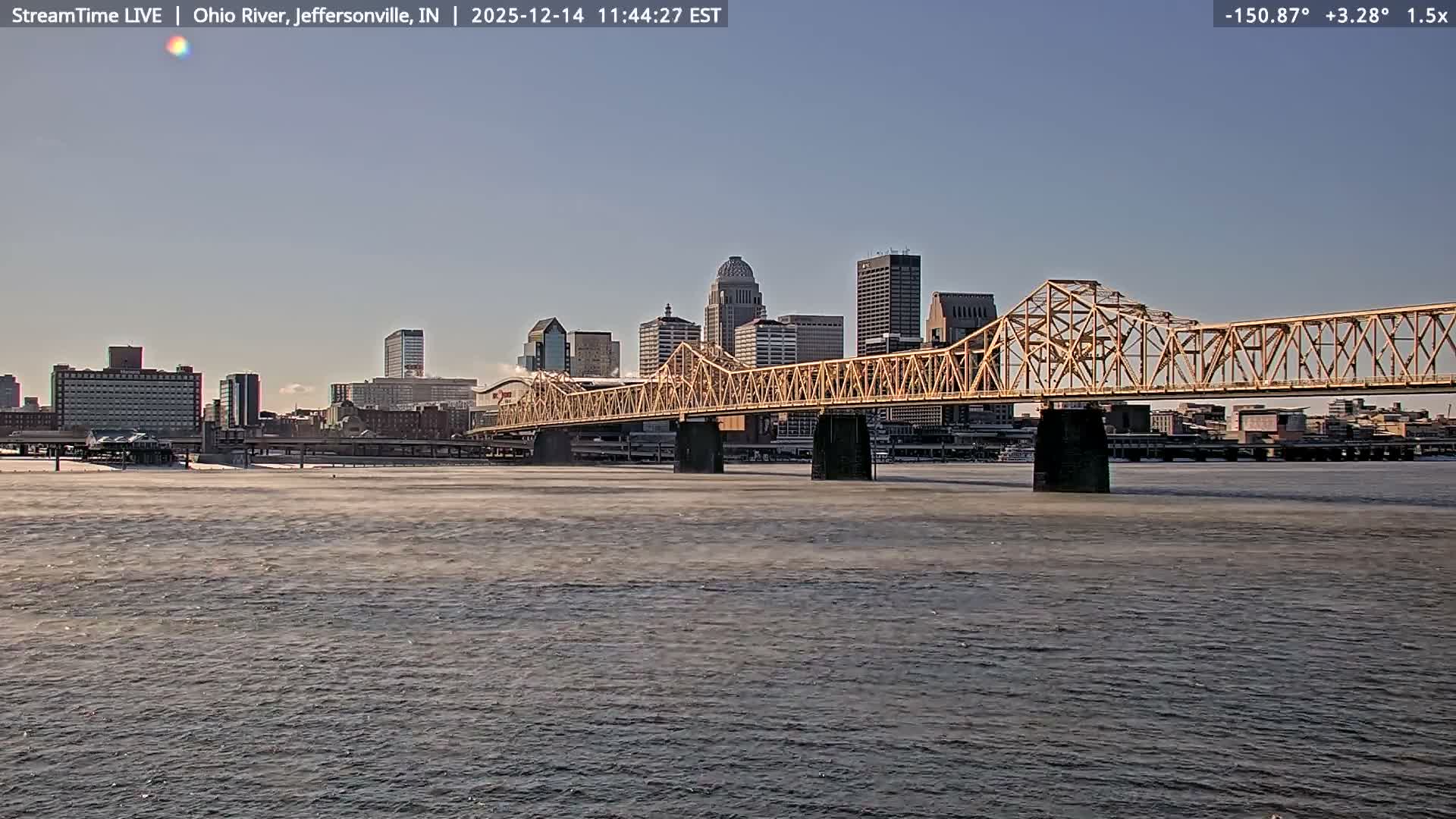 A cloudy nighttime scene reveals a distant city skyline brightly lit with various colored lights and a large illuminated truss bridge spanning a wide river, all reflecting on the dark water.