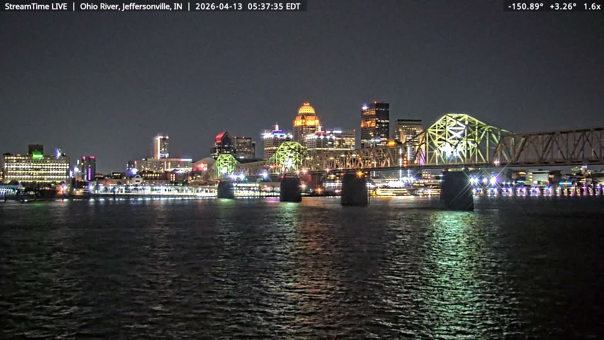 A cloudy nighttime scene reveals a distant city skyline brightly lit with various colored lights and a large illuminated truss bridge spanning a wide river, all reflecting on the dark water.
