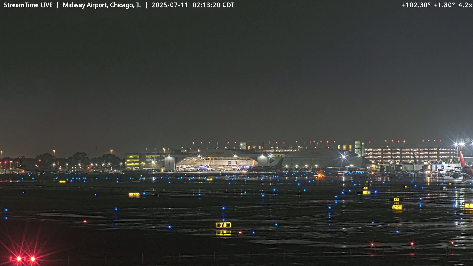 A nighttime view of an airport tarmac with numerous lights illuminating the wet ground and airplanes near a brightly lit terminal building under a dark sky.