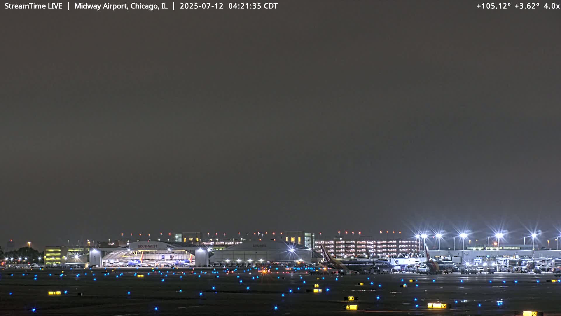 A nighttime view of an airport tarmac with numerous lights illuminating the wet ground and airplanes near a brightly lit terminal building under a dark sky.