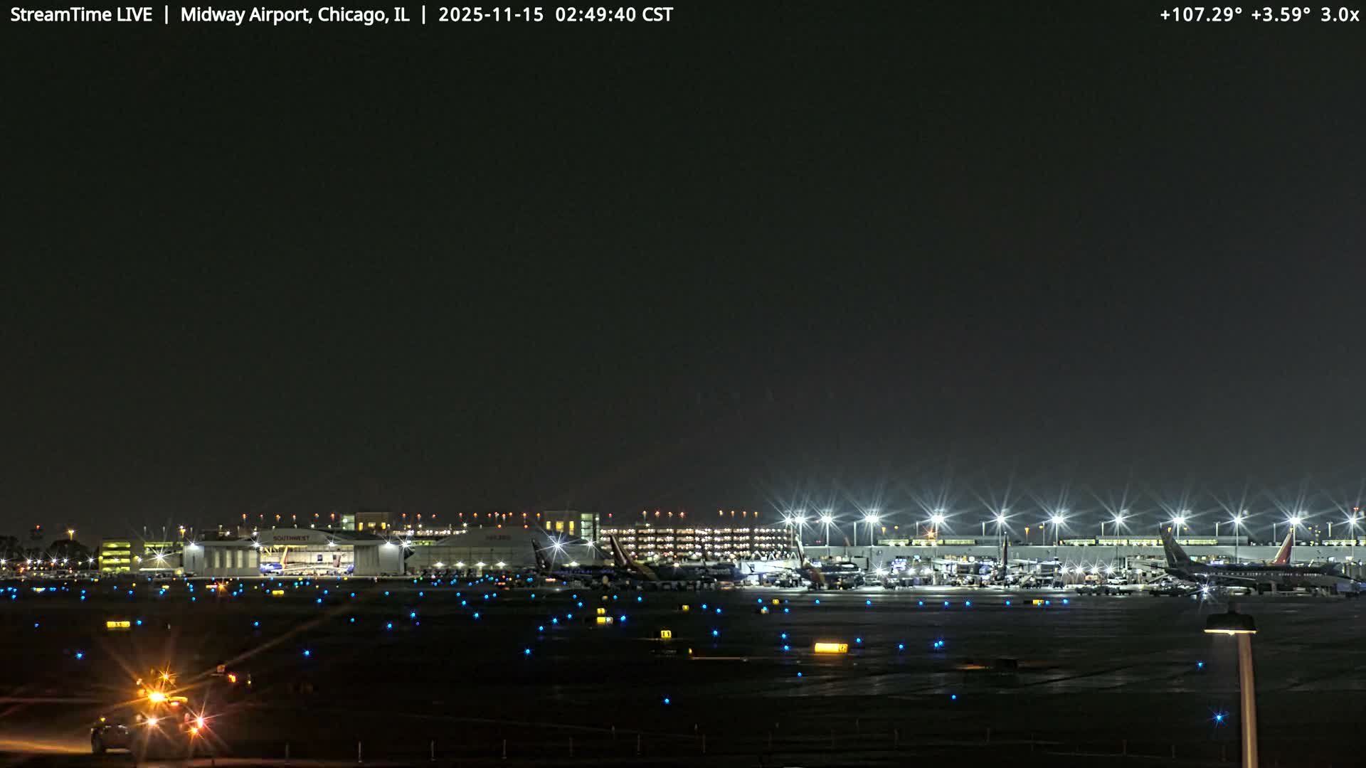 A wide view of an airport at night shows multiple airplanes parked at brightly lit terminals and hangars, with illuminated blue taxiway lights across the tarmac and a service vehicle in the foreground, all under a clear, dark sky.