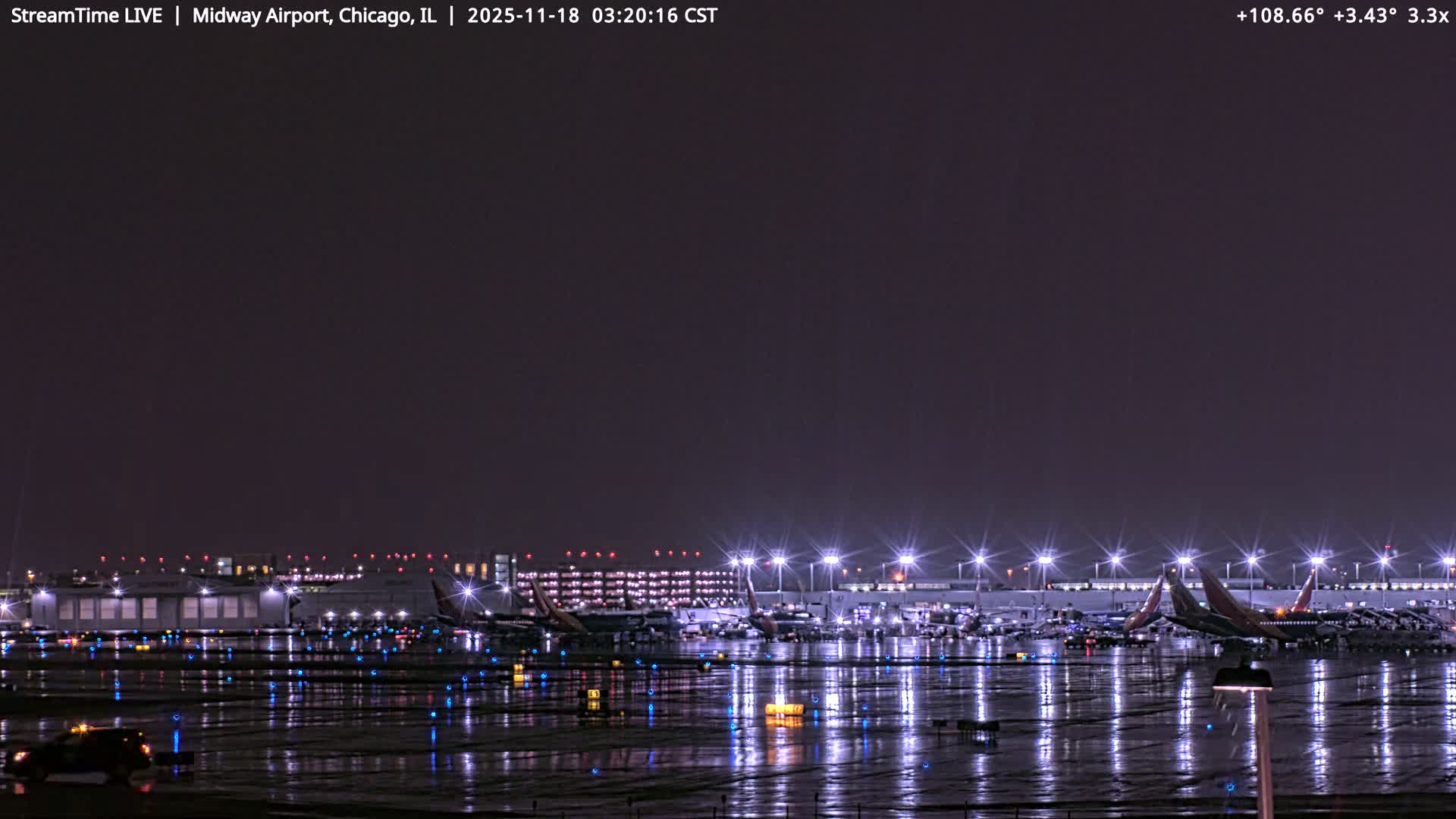 A wide view of an airport at night shows multiple airplanes parked at brightly lit terminals and hangars, with illuminated blue taxiway lights across the tarmac and a service vehicle in the foreground, all under a clear, dark sky.