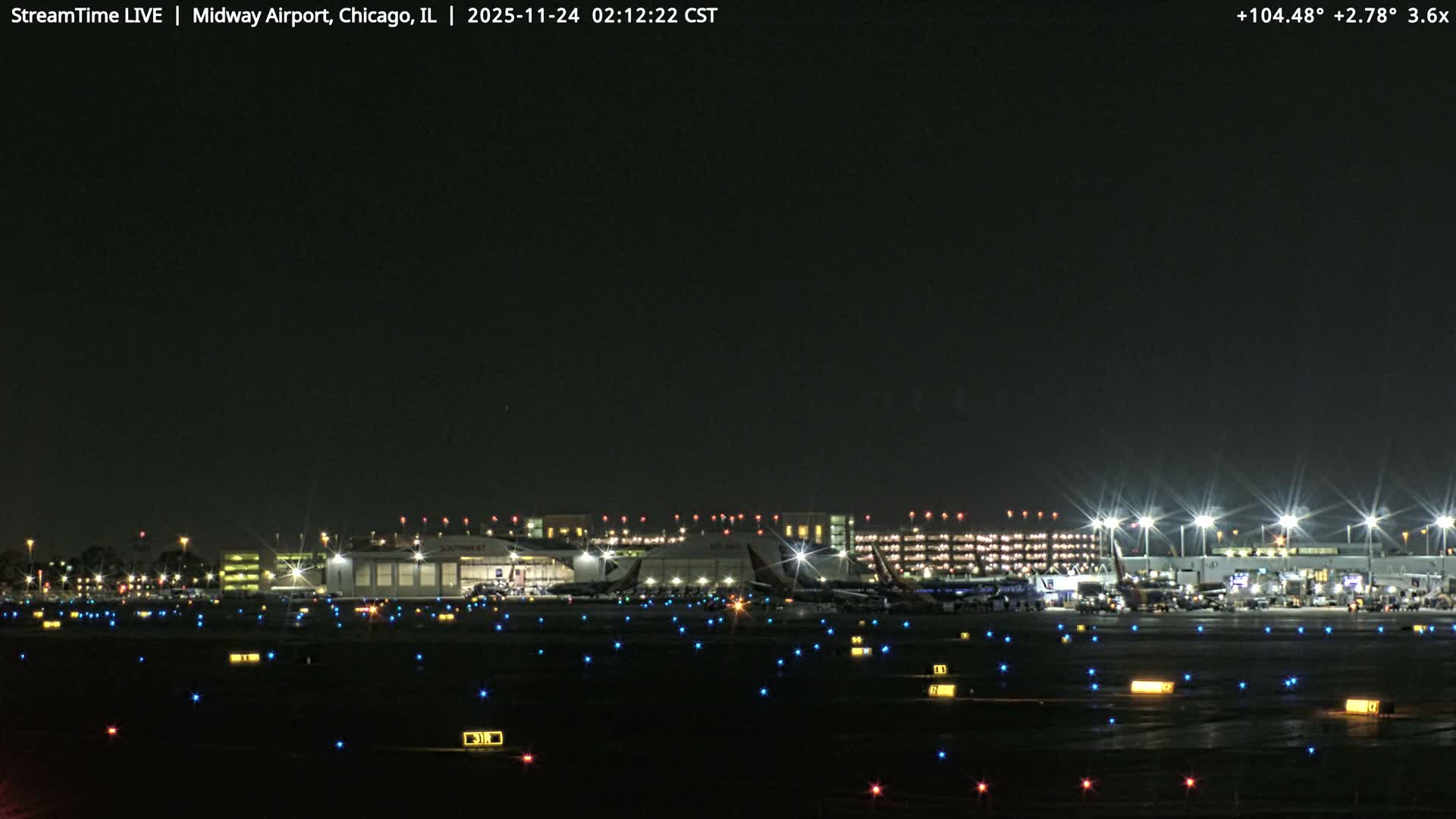 The image displays an airport tarmac and terminal buildings brightly lit at night under clear skies, with multiple parked aircraft and an array of blue, red, and yellow ground lights marking the taxiways.