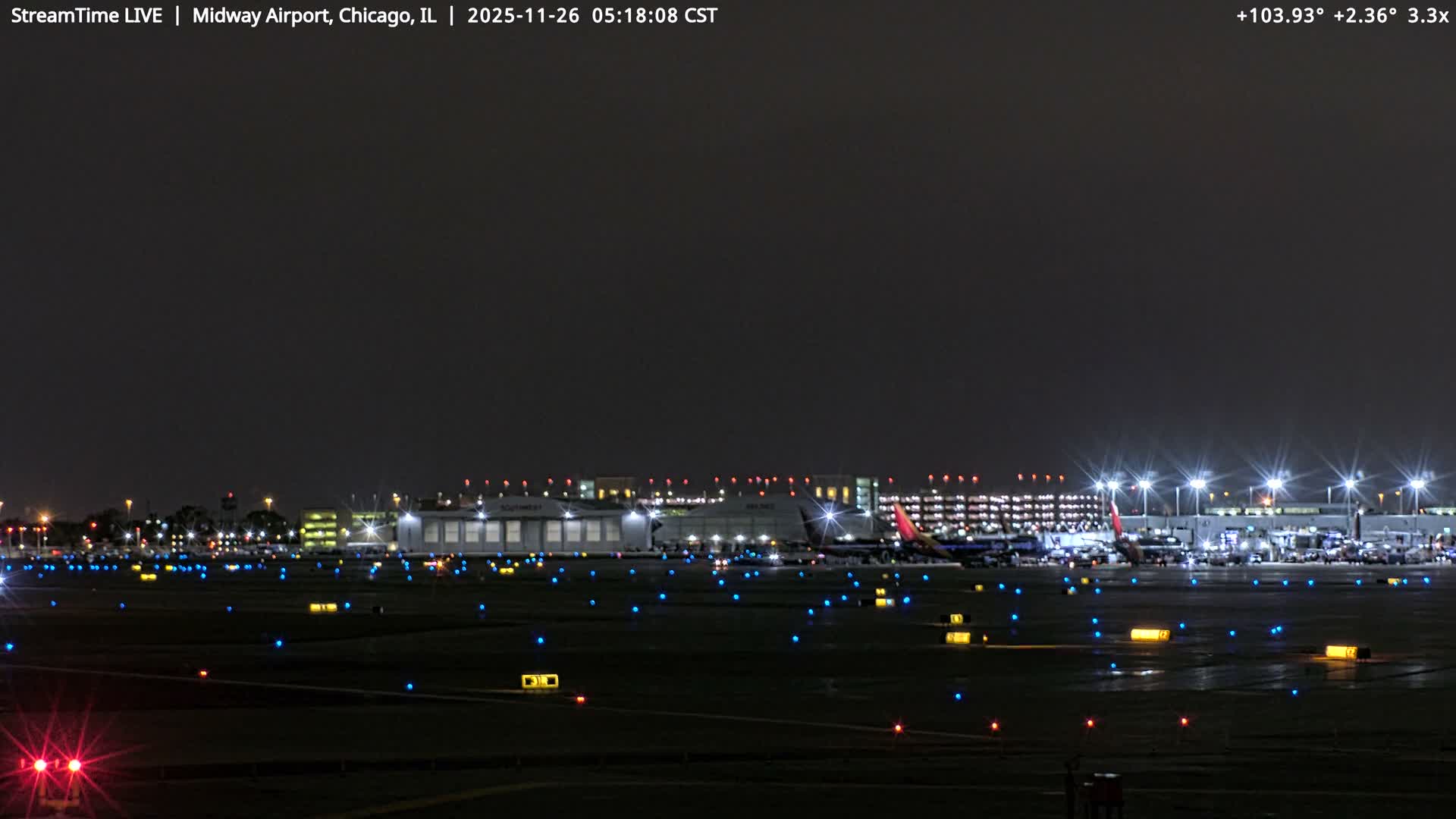 The image displays an airport tarmac and terminal buildings brightly lit at night under clear skies, with multiple parked aircraft and an array of blue, red, and yellow ground lights marking the taxiways.