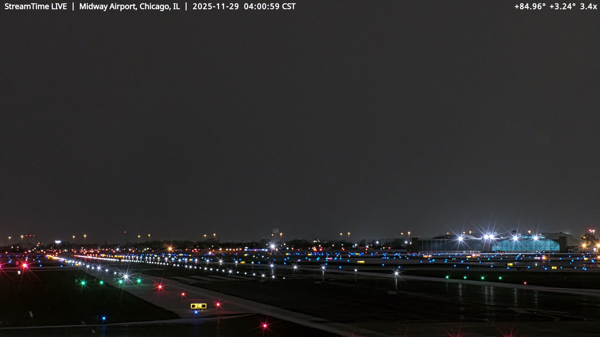The image displays an airport runway at night, vividly illuminated by an array of red, green, blue, and white lights under a clear, dark sky, with distant airport structures and city lights visible on the horizon.