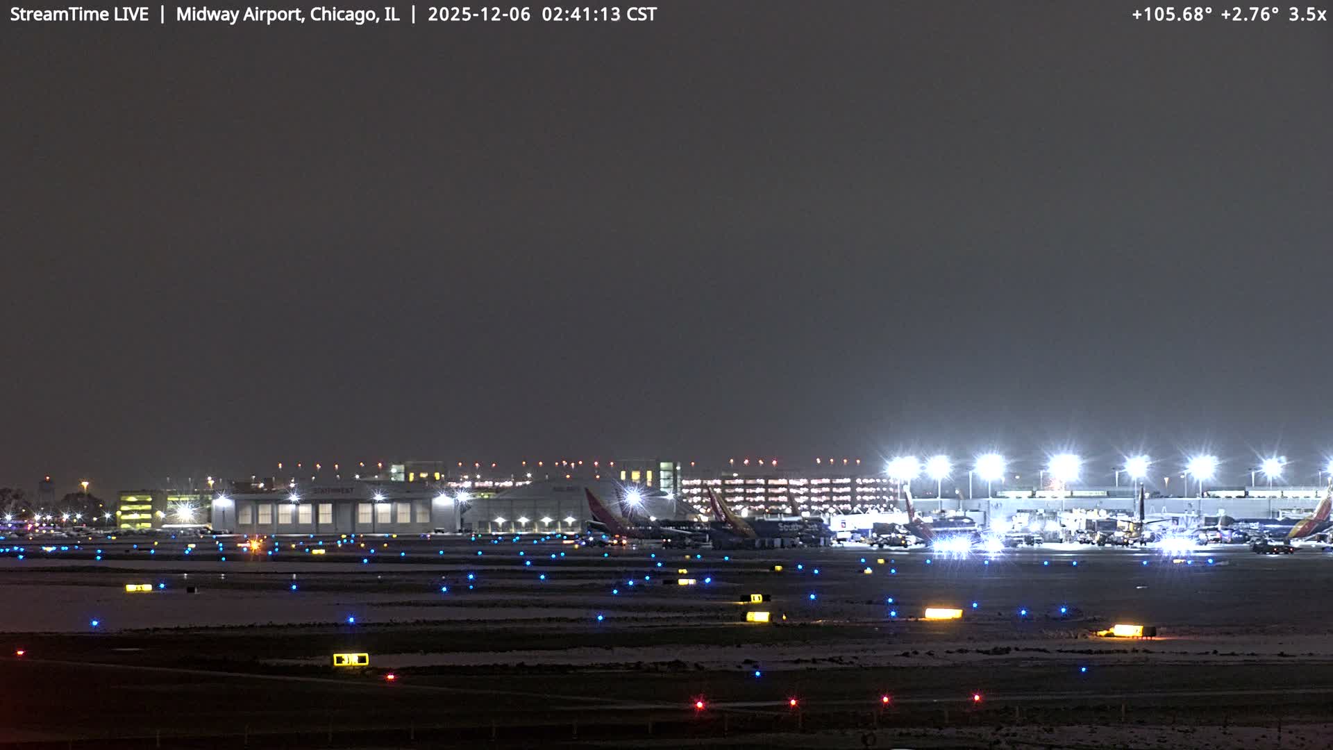 A nighttime view of an airport, featuring multiple parked airplanes, brightly lit terminals and taxiways with blue, yellow, and red lights, and a light dusting of snow on the ground under a dark, overcast sky.