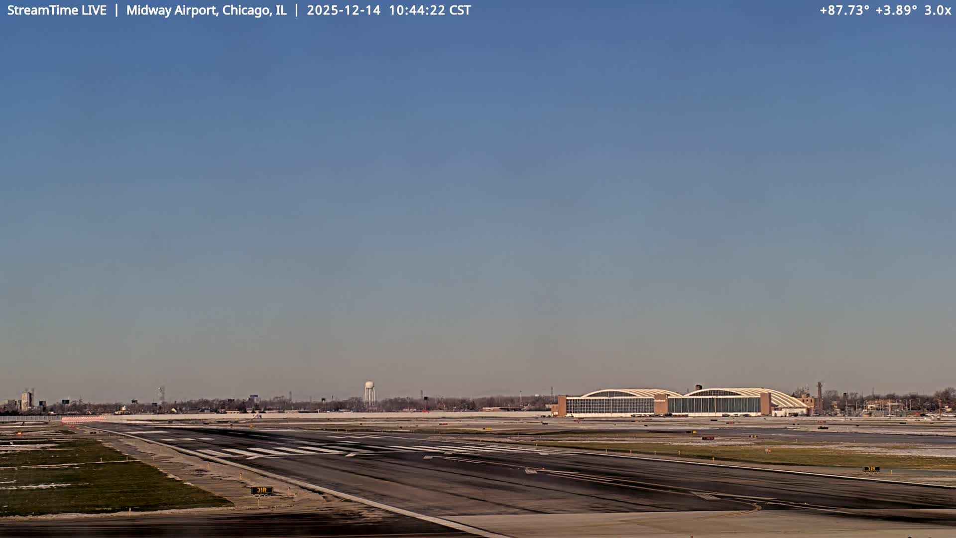 A nighttime view of an airport, featuring multiple parked airplanes, brightly lit terminals and taxiways with blue, yellow, and red lights, and a light dusting of snow on the ground under a dark, overcast sky.