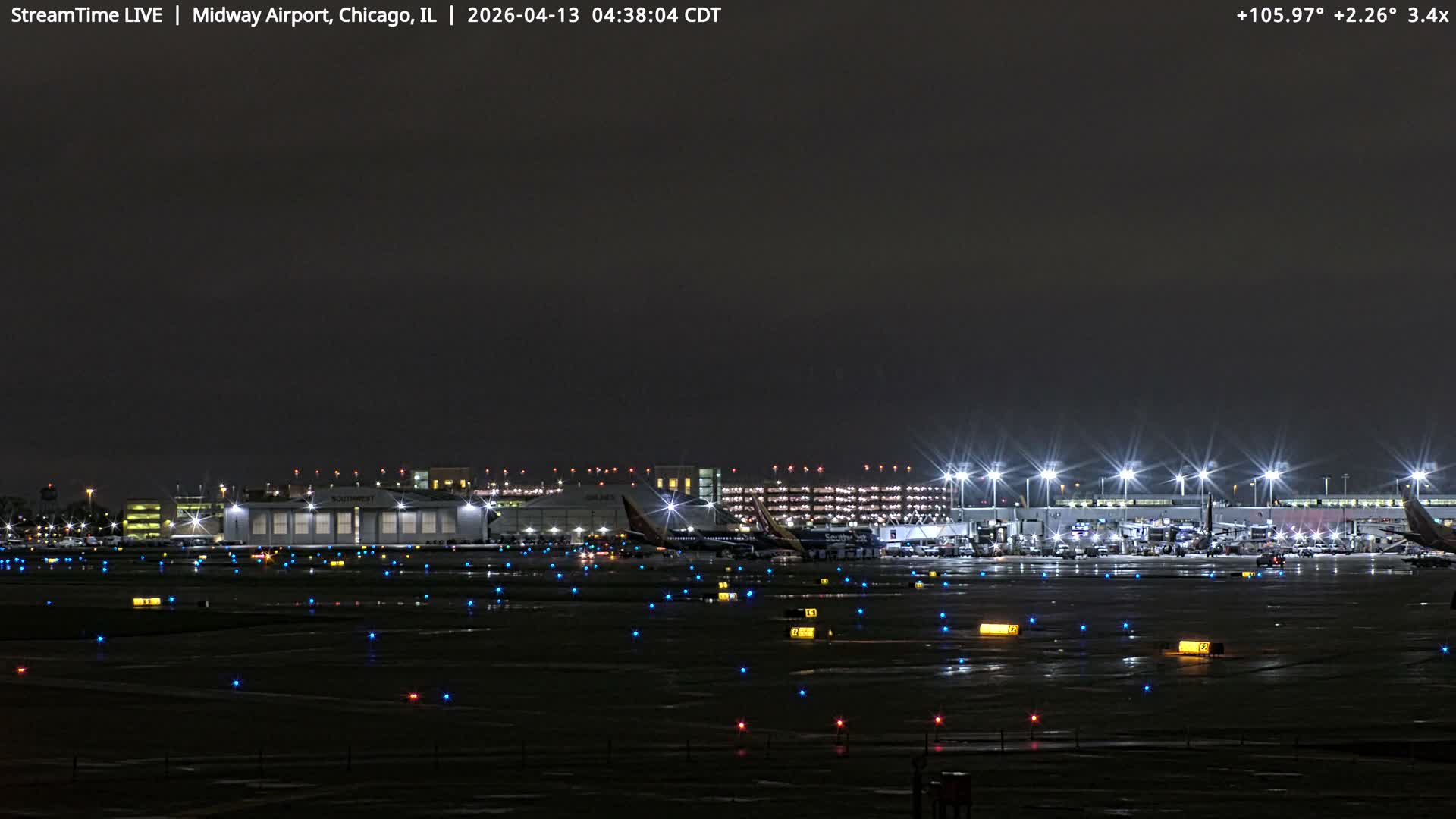 A nighttime view of an airport, featuring multiple parked airplanes, brightly lit terminals and taxiways with blue, yellow, and red lights, and a light dusting of snow on the ground under a dark, overcast sky.